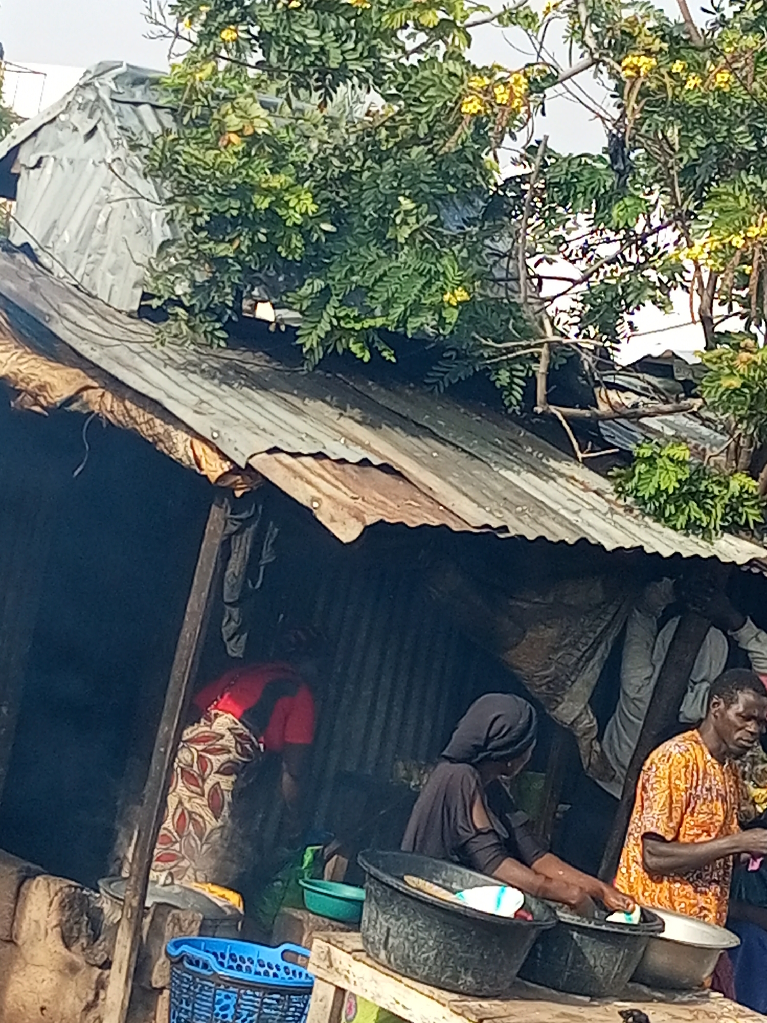 This image depicts a scene in the city of Tumfure, Nigeria. In the foreground, a person wearing a patterned skirt and a red top is standing and appears to be engaged in some activity. To their right, a person wearing a black headscarf and a black top is actively washing dishes in a large black basin. Next to them, a person in an orange shirt is also tending to dishes. In front of these individuals, a blue plastic basket is visible. The background reveals a makeshift structure with a corrugated metal roof, partially obscured by lush green foliage and yellow flowers from an overhead tree. The overall environment suggests a functional, albeit rustic, outdoor setting likely associated with domestic activities or a marketplace.