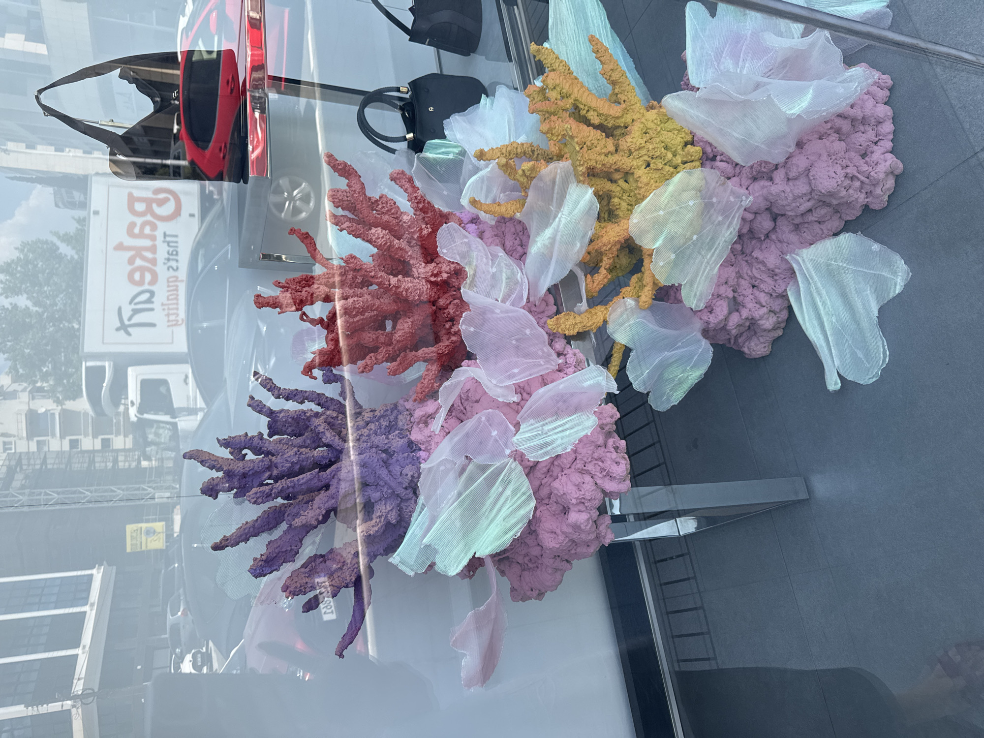 The image captures a vibrant storefront display, likely in Limassol, Cyprus, featuring an artistic arrangement of colorful, coral-like structures. The display is set against a backdrop of reflections from the street, showing buildings, vehicles, and a truck with Cyrillic lettering that reads "BAKERY." The foreground is dominated by the artificial coral, rendered in shades of purple, pink, red, and yellow, interspersed with translucent, iridescent fabric pieces resembling sea anemones or jellyfish. These elements are placed on a textured pink base, supported by a sleek, mirrored table.
To the right of the coral display, two black handbags are visible on a shelf, one with a gold clasp bearing the letter "A." The store window itself reflects the urban environment, suggesting a commercial area. The floor tiles are dark grey, with a subtle pattern. While no people are clearly depicted as being inside the store, a sliver of a person's leg and foot wearing sandals is visible in the bottom left corner of the frame, indicating a passerby's presence outside the window. The overall impression is a stylized, underwater-themed display designed to attract attention.