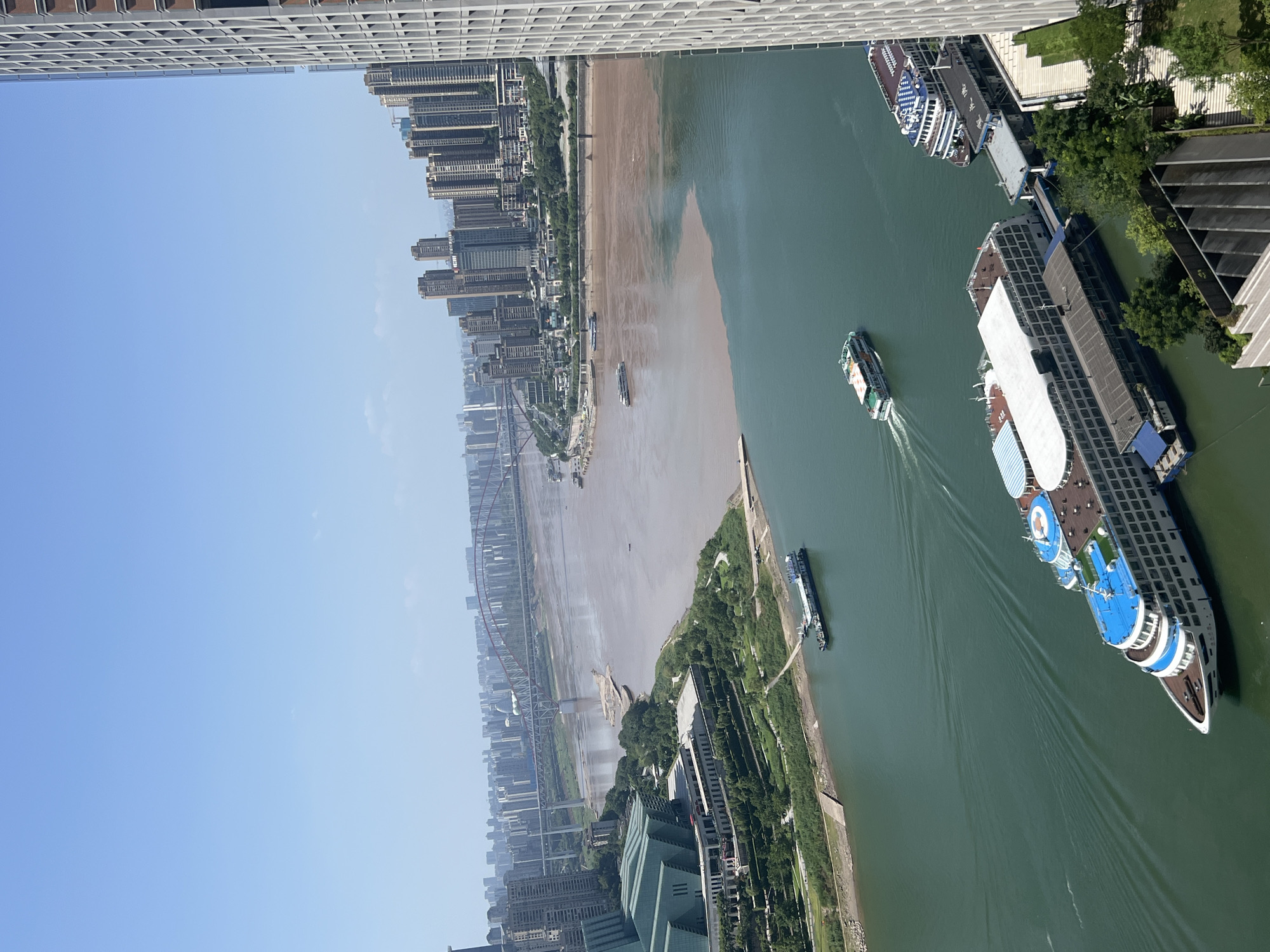 This aerial image captures the confluence of two rivers in the Yuzhong District, Chongqing, China. The left bank features a modern green-roofed building with tiered landscaping and walkways. Several boats are visible on the water, including a large white and blue cruise ship docked on the right side of the frame. In the distance, multiple bridges span the river, connecting a sprawling cityscape dominated by high-rise buildings. The water itself shows a distinct color separation, with one section appearing muddy brown and the other a clear green. The sky above is clear blue with a few wispy clouds. The scene is brightly lit, suggesting it is daytime.