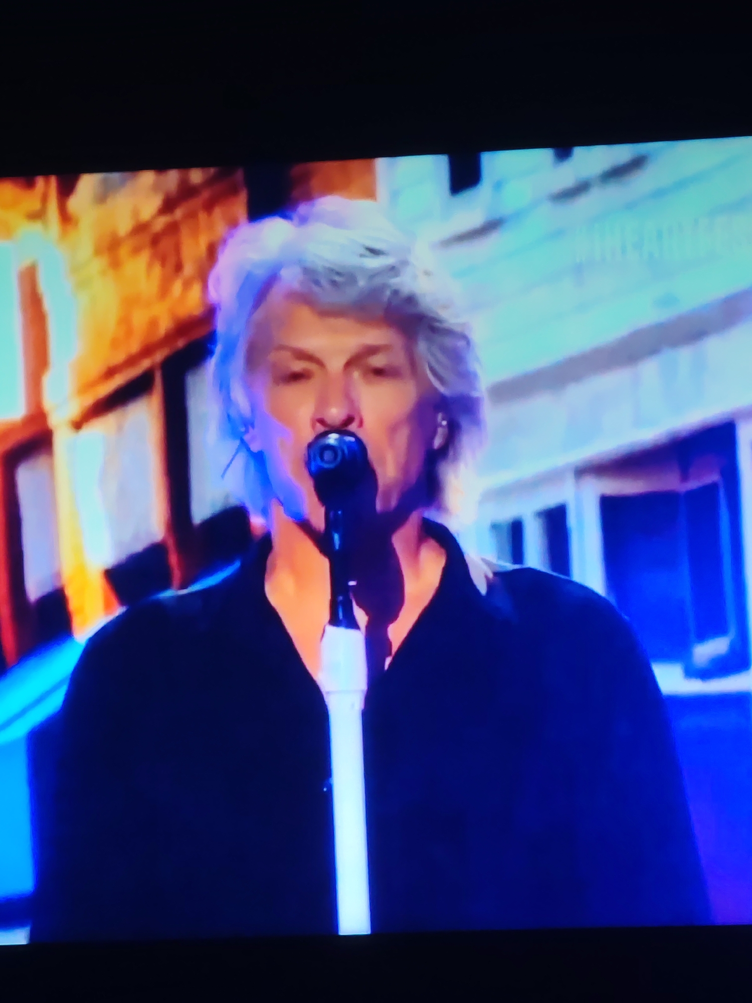 The image shows a medium shot of a man with graying, voluminous hair, wearing a black collared shirt. He is holding a microphone to his mouth, suggesting he is singing or speaking. The background is blurry and appears to be a stage set with abstract colorful elements and what looks like a structure resembling a building facade. There are no discernible elements in the image that would allow for the identification of the city of Vladimir, Russia, or any specific location within it.