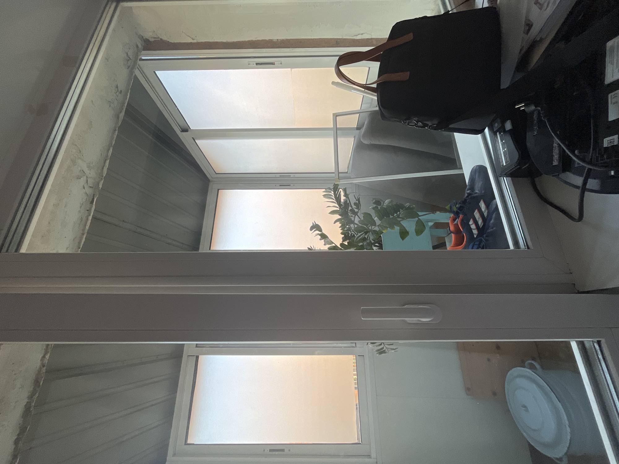 The image shows an interior view of a room, possibly a balcony or enclosed patio, with large windows. The windows are frosted and allow a soft, warm light to filter through, suggesting either sunrise or sunset. Through the windows, a hazy outdoor scene is visible, with hints of buildings. Inside the room, on a windowsill, are several objects. A black tote bag with brown leather handles sits prominently on the right. To its left, a pair of navy blue athletic shoes with white and orange accents are visible. In the background, to the left, a large light blue plastic tub with a lid is partially seen, resting on a wooden surface. A green leafy plant in a teal pot is also visible. The ceiling appears to be unfinished concrete with corrugated metal panels. The overall impression is one of a functional, somewhat cluttered indoor space adjacent to an outdoor area.
