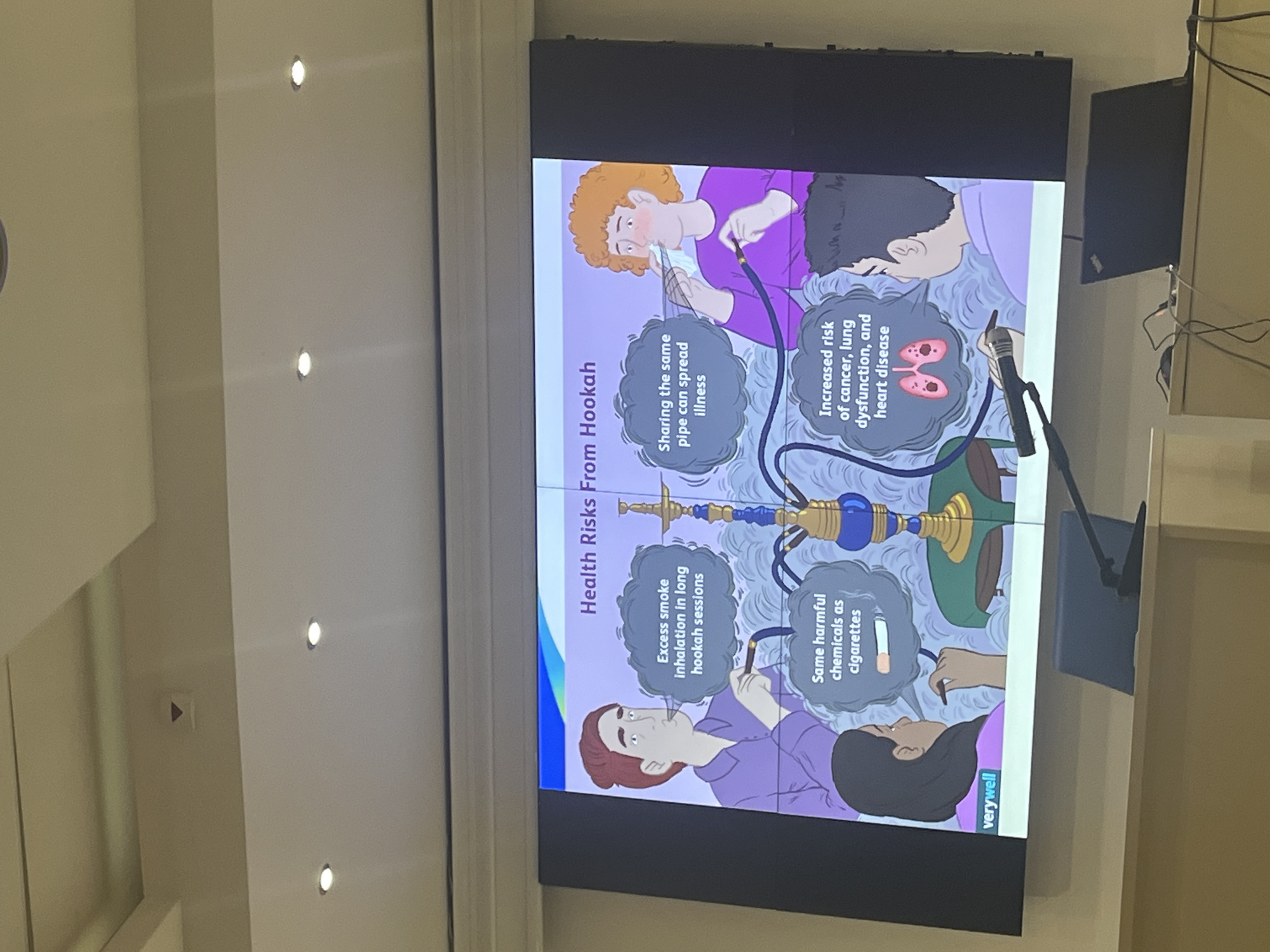 The image displays a presentation screen in an indoor setting, likely a conference or lecture hall, situated in Plovdiv, Bulgaria. The large screen, composed of multiple panels, is showing a vibrant graphic titled "Health Risks From Hookah."
The graphic features illustrations of four individuals—two men and two women—all depicted with a hookah. A stylized hookah with a blue base and golden elements is centrally positioned. Surrounding the figures are text bubbles outlining various health hazards associated with hookah use.
Visible text on the graphic includes:
- The main title: "Health Risks From Hookah."
- "Sharing the same pipe can spread illness," accompanied by an illustration of a person holding the pipe.
- "Excess smoke inhalation in long hookah sessions," with an illustration of a person exhaling smoke.
- "Same harmful chemicals as cigarettes," featuring an image of a cigarette and a person inhaling.
- "Increased risk of cancer, lung dysfunction, and heart disease," alongside stylized depictions of lungs and a heart.
The "verywell" logo or credit is visible at the bottom of the graphic.
In front of the screen, a microphone is partially visible, suggesting that a presentation or lecture is taking place. Cables are also seen on the right side of the frame, connected to unseen electronic equipment. The surrounding walls are light-colored, and several recessed spotlights illuminate the ceiling. The image, which is rotated ninety degrees clockwise, captures an educational or public health awareness event.