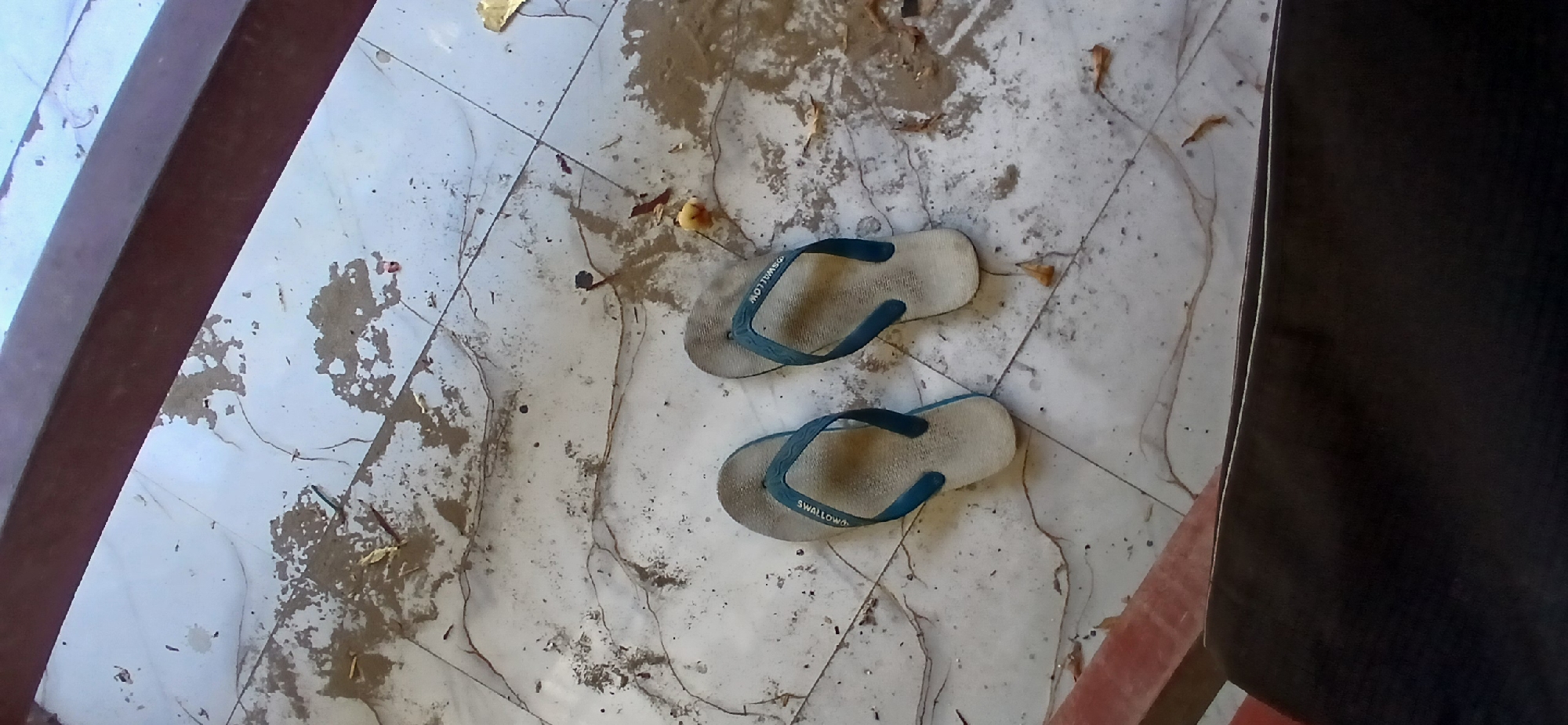 On a dirty, tiled floor, a pair of blue and white flip-flops are placed. The tiles appear to be light-colored with gray and brown stains and streaks, suggesting they have not been cleaned recently. There are also some scattered leaves and debris on the floor. The flip-flops are positioned side-by-side, with the right flip-flop slightly in front of the left. The straps of the flip-flops are blue, and the soles are white with a textured pattern. The word "SWALLOW" is visible on the sole of the right flip-flop, and a similar marking is likely on the left one. The scene appears to be outdoors, possibly on a porch or patio, given the natural light and the presence of debris. The setting is in Pejaten, Indonesia.
