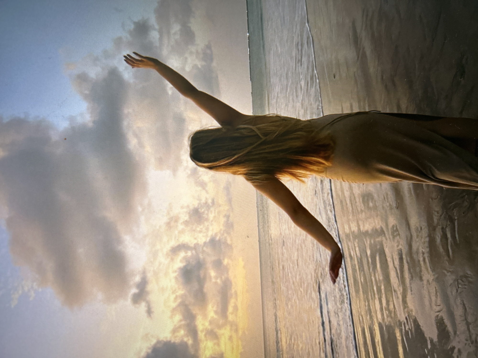 A woman stands on the beach, bathed in the light of a Kuta Selatan, Indonesia sunset. Her back is to the viewer, with both arms extended upward. Her long blonde hair flows down her back as she gazes up to the horizon. She is wearing a flowing, light-colored dress. The sky is filled with clouds, illuminated by the setting sun. The beach is reflective and light-colored. The ocean edge is visible in the background.