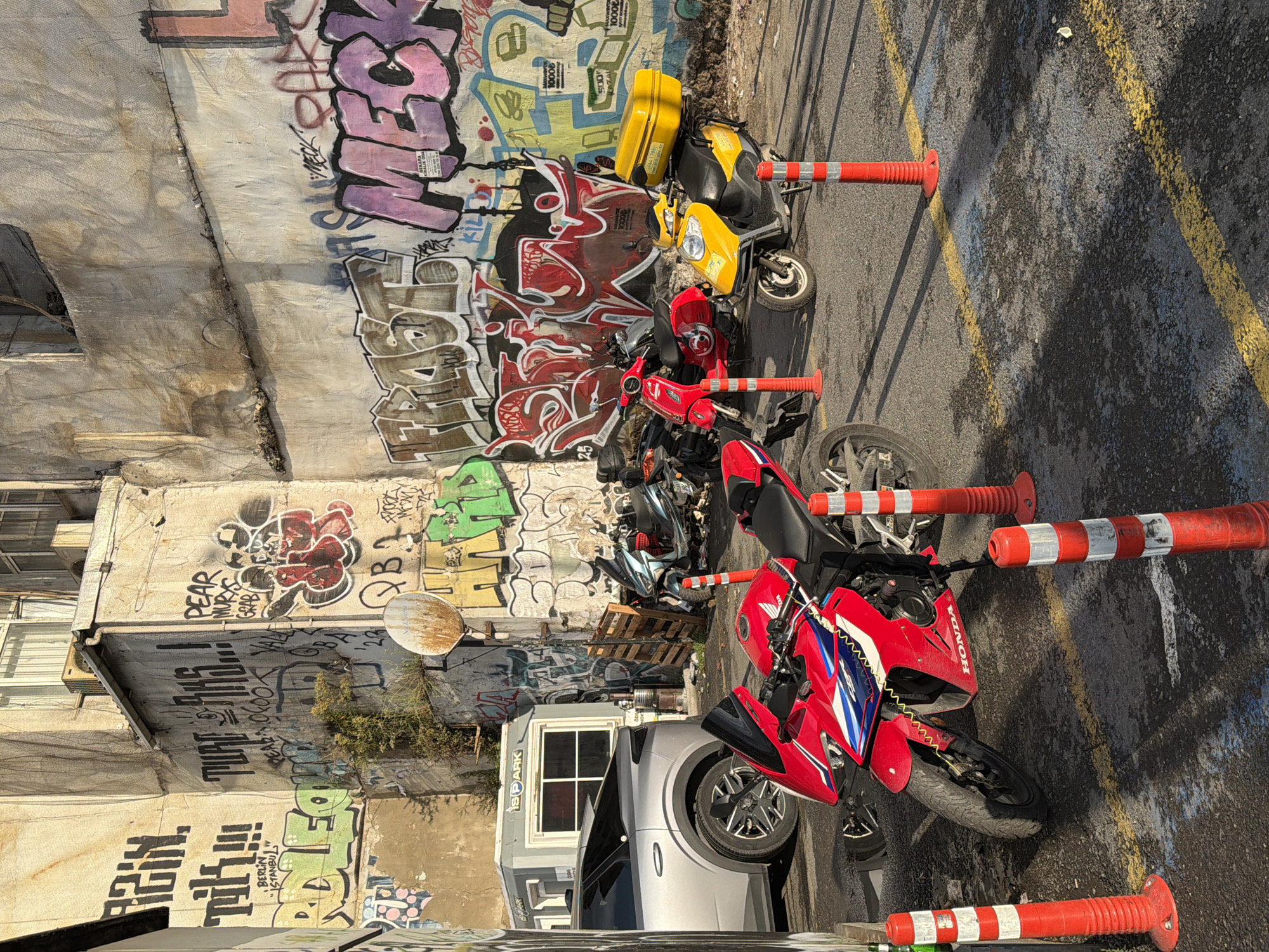 In the Galata district of Beyoğlu, Turkey, an outdoor scene is depicted, featuring a collection of motorcycles and a car parked against a wall adorned with graffiti. A red Honda motorcycle is prominently positioned in the foreground. Behind it, a yellow scooter with a top box is parked, along with several other motorcycles and scooters. A silver car is visible on the left side of the image. The environment is characterized by a weathered wall covered in vibrant graffiti, a small portable structure with a sign reading "SPARK", and several red and white striped traffic cones. The ground is asphalt, marked with yellow lines, and shows signs of dampness and scattered debris. The lighting suggests it is daytime, with shadows cast by the vehicles and cones.