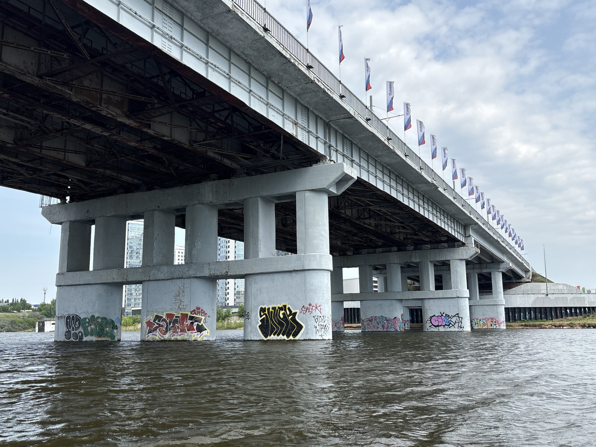The image depicts the underside of a concrete and steel bridge spanning a body of water. The bridge supports are adorned with graffiti. A line of flags is visible along the bridge's edge. In the background, there is a skyline with buildings. The scene is located in Naberezhnye Chelny, Russia.