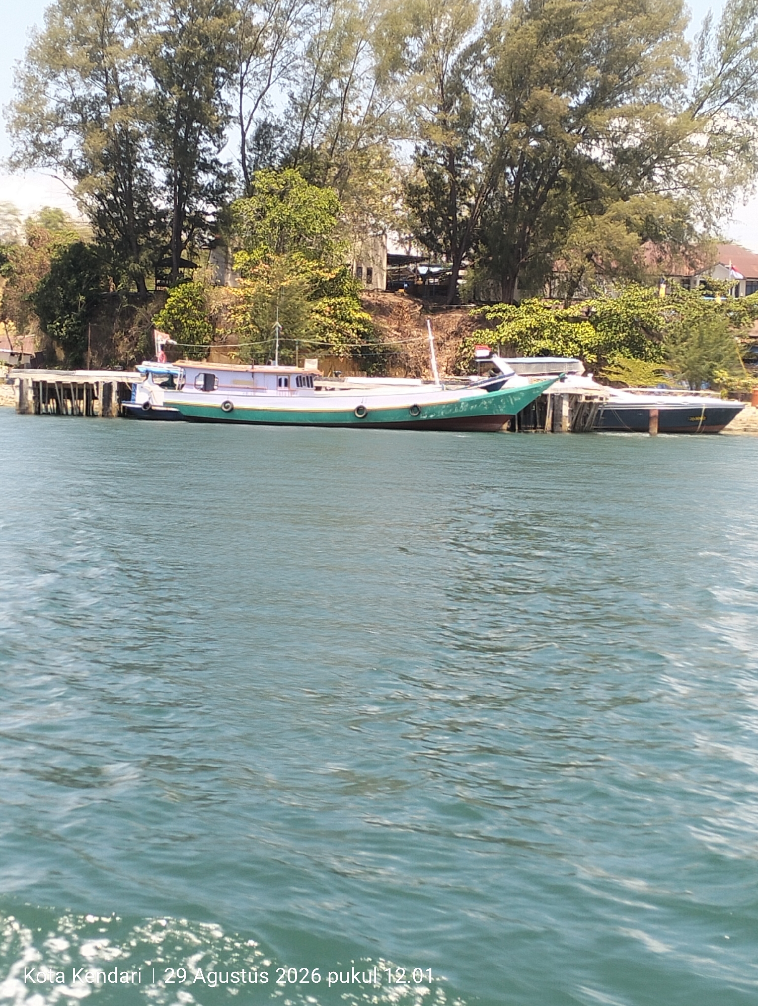 An eye-level, medium shot captures a partially submerged longboat docked alongside a pier on a body of calm, blue-green water. The boat, painted with a prominent green stripe along its waterline and a lighter colored upper hull, is moored to a wooden pier that extends from a densely vegetated shoreline. The shore is lined with tall trees and some low-lying shrubs. Buildings are visible through the foliage on the higher ground behind the trees. The water exhibits small ripples and reflections, indicating a bright day. In the foreground, the water is textured with white foam, suggesting the viewer is on another vessel. Text overlaid on the bottom left corner of the image reads "Kota Kendari | 29 Agustus 2026 pukul 12.01".