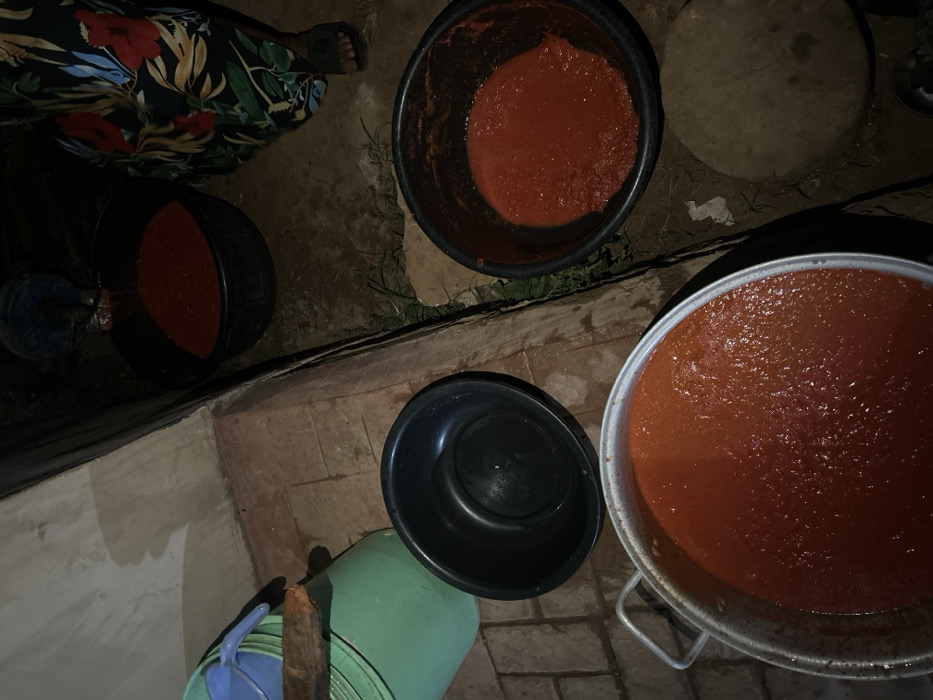 Four buckets and a large pot filled with red sauce are arranged on a brick surface, outside near a grassy area. One bucket is empty, sitting atop a green plastic container. The sauce appears consistent in each container. A person's legs and patterned clothing are visible in the top left corner. The setting is dimly lit.