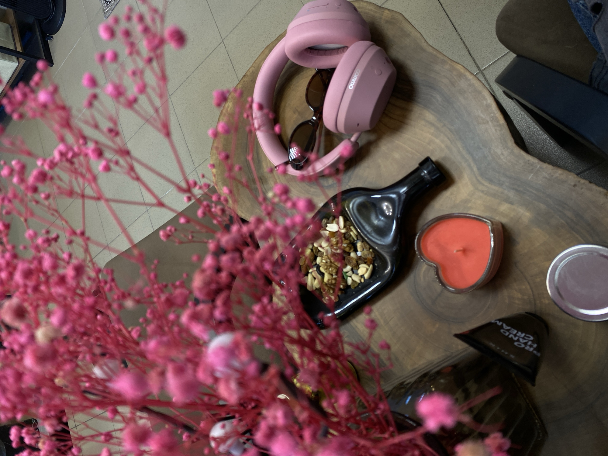 The image depicts a still life arrangement on a wooden surface. Dominating the foreground are pink gypsophila flowers, blurring some of the details behind. In the upper-right quadrant are pink headphones with sunglasses tucked inside. Adjacent to the headphones is a black, bottle-shaped dish containing a mix of nuts and dried fruit. A heart-shaped candle sits next to the dish. Further right, there is a round container and a tube of hand cream. The arrangement appears to be on a coffee table next to a seat. The background reveals a tiled floor. The location is not discernible from the image, so the city of Votkinsk, Russia, cannot be confirmed.