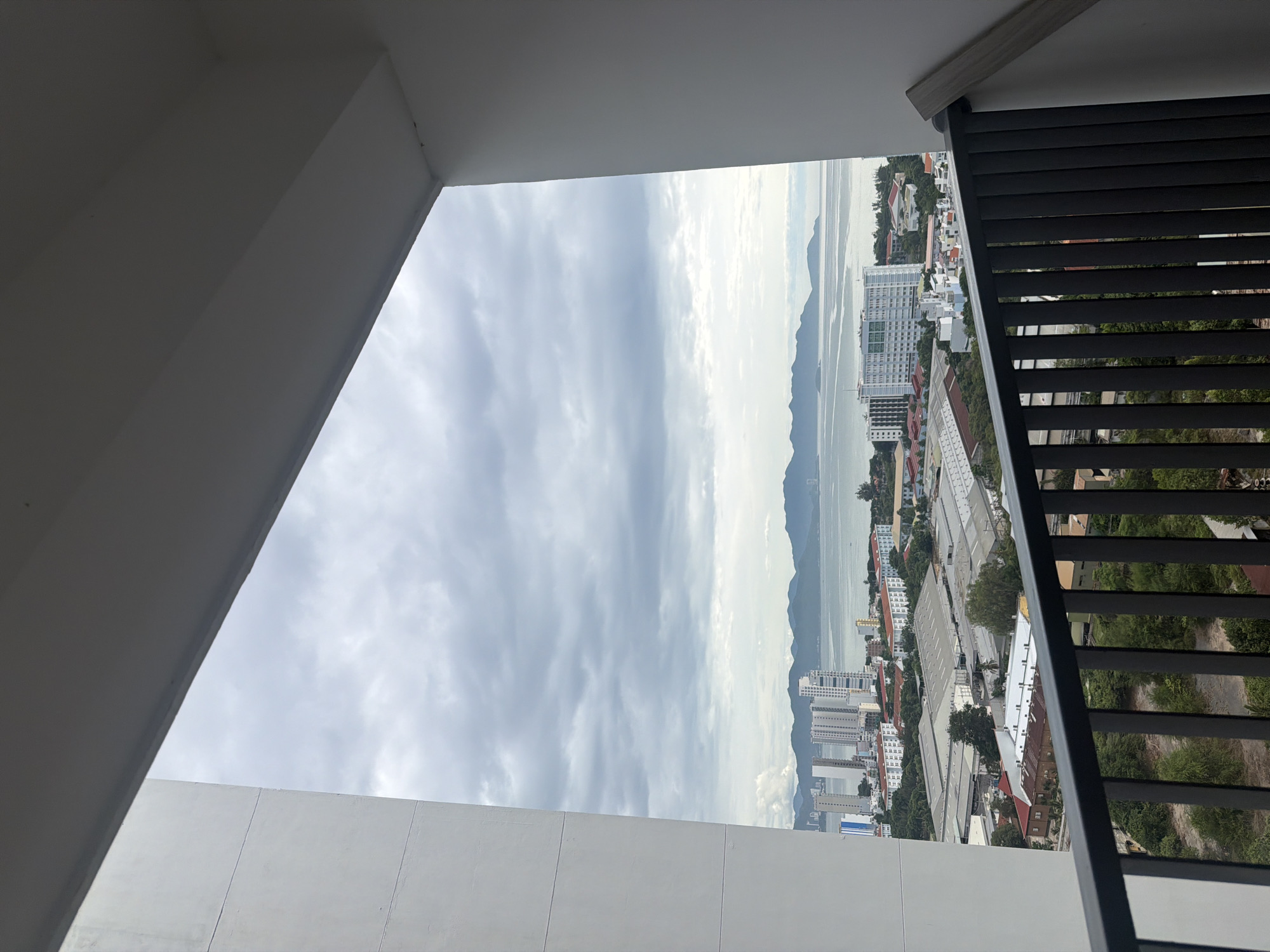 This is a slightly elevated, wide-angle shot taken from a balcony overlooking a coastal city. The foreground consists of a dark gray metal railing. Beyond the railing, the scene opens up to reveal a densely populated urban area. Many buildings of varying heights, including several tall high-rises, are visible. These structures are predominantly white or light-colored, with some displaying red-tiled roofs. In the middle ground, a large industrial complex with multiple warehouse-style buildings featuring light-colored roofs is present. The background features a large body of water, likely the sea, with a range of hills or mountains on the horizon. The sky is overcast with gray clouds. The overall impression is a view of an urban coastal landscape under a cloudy sky. The presence of high-rise buildings, industrial complexes, and the sea suggests a coastal city in Southeast Asia. The style of architecture and the surrounding landscape are consistent with Nha Trang, Vietnam. There are no people visible in the image.