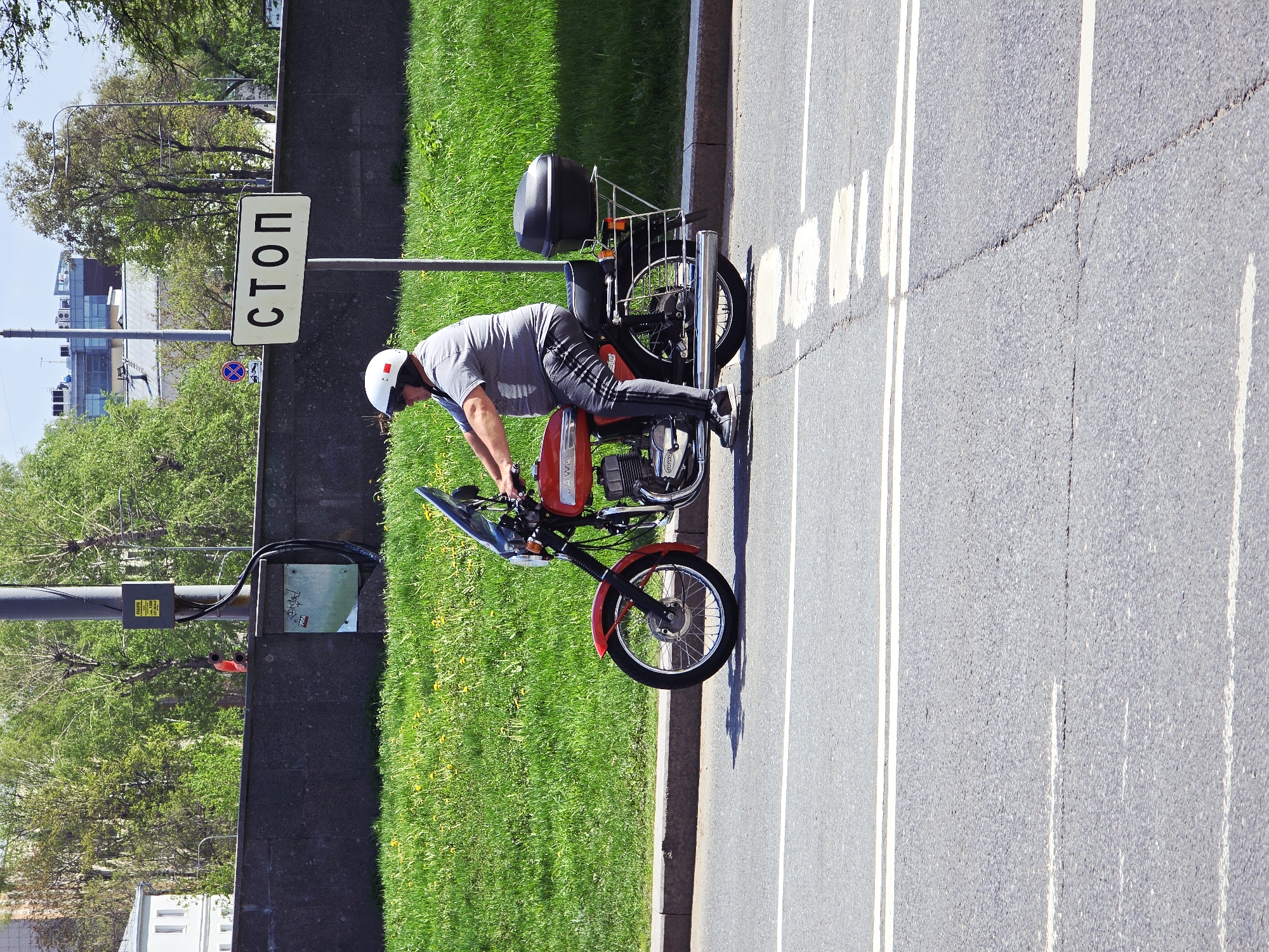 The image shows a man on a red Jawa motorcycle stopped on the side of a road, next to a grassy area, with a "СТОП" (STOP) sign visible. The location appears to be Moscow, Russia, based on the Cyrillic alphabet sign and architectural features. The man is wearing a white helmet, a gray t-shirt, and patterned pants. He is leaning forward on the motorcycle. The road is marked with white lines. The motorcycle has a black storage box mounted behind the seat. There is a utility pole and other signage nearby. The background includes buildings and trees, indicating an urban or suburban setting.