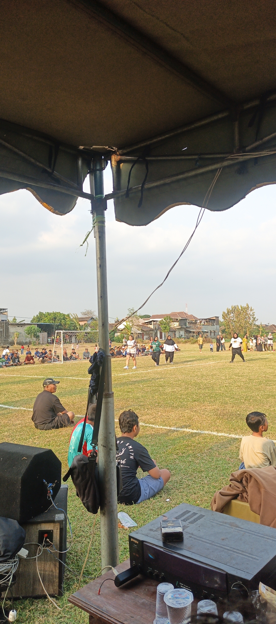 This is a vertical, eye-level, outdoor medium shot of a soccer game in progress on a grass field, framed by a tent canopy in the foreground. The game appears to be between two women's teams. On the field, one woman in a white and black uniform is running, while another woman in a green and black hijab is walking. Several spectators are seated on the grass, some close to the camera and others further back near a soccer goal. A speaker system and karaoke machine are set up on a wooden table in the immediate foreground, with wires connected. The background shows a line of residential buildings and trees under a hazy sky. The lighting suggests it is late afternoon.
