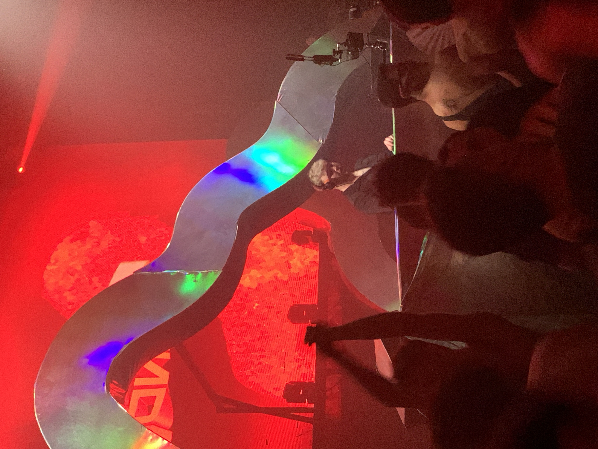 The image depicts a dimly lit nightclub or concert venue, likely in Tel Aviv, Israel, due to the presence of a visible "AV" logo in the background, which is associated with the AV club in Tel Aviv. The scene is dominated by red lighting and a large, abstract, iridescent structure that curves across the upper portion of the frame.
In the foreground, a crowd of people is visible, silhouetted against the stage lighting. Many are raising their hands, suggesting they are engaged with the performance.
On the stage, a DJ wearing headphones is at work. They are dressed in a dark shirt and appear focused on their equipment. Behind the DJ, a large screen displays a red and orange abstract graphic. Several speakers are also visible on the stage.
The overall atmosphere is energetic and immersive, characteristic of a live music event. The iridescent structure and the vibrant red lighting contribute to a modern and dynamic visual aesthetic.
