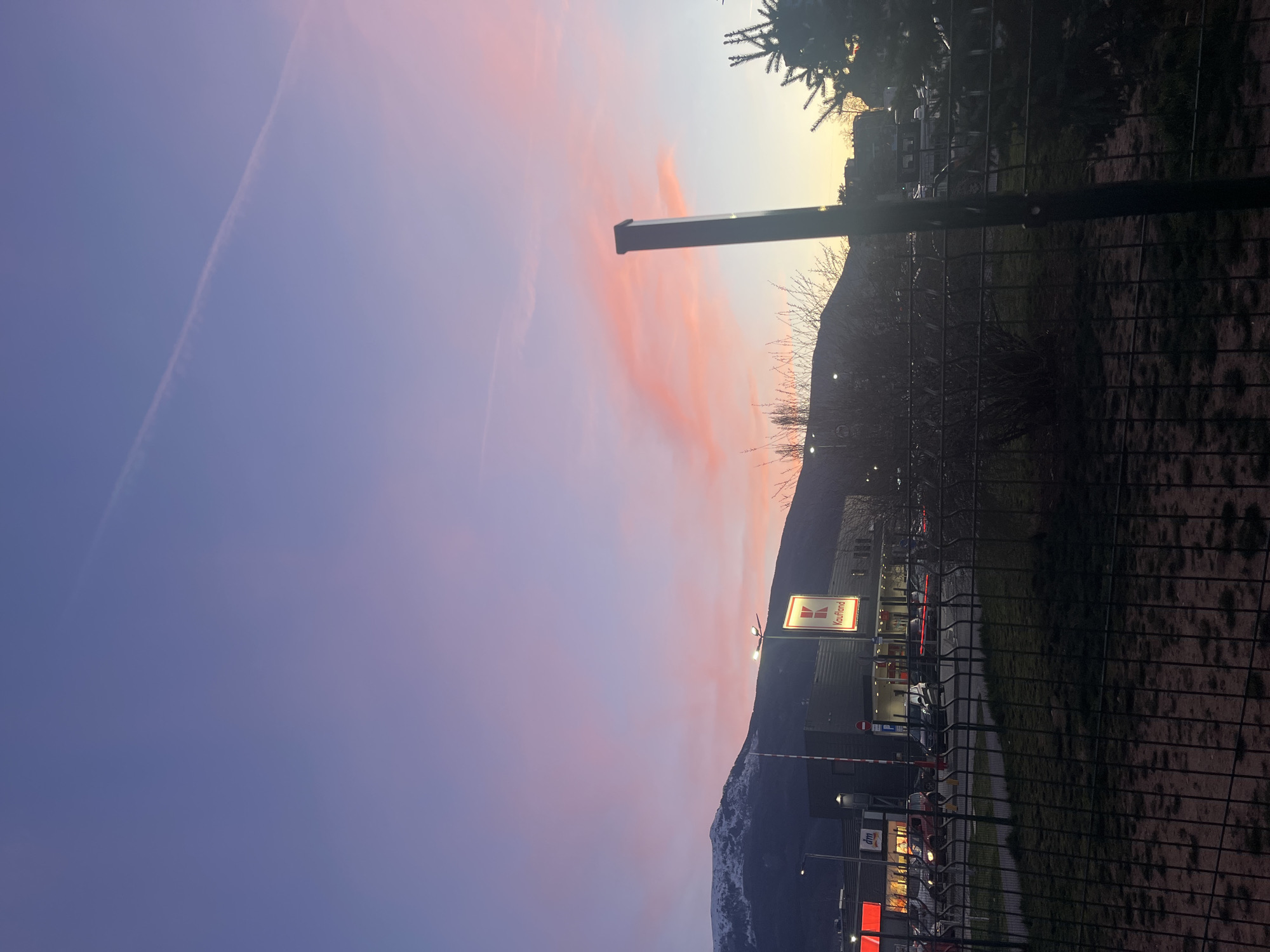 The image shows a sunset sky over a cityscape, likely taken near a Kaufland store. The sky features a gradient of colors, from pale orange near the horizon to purple in the upper portion, with some pink clouds. In the foreground, there's a fence and what appears to be a pole. The Kaufland store's sign is visible, along with other buildings and parking areas. Mountains are present in the middle ground, partially covered with snow. The scene is illuminated by the setting sun and artificial lights. It's not possible to confirm the exact location as Sofia, Bulgaria from the image alone.