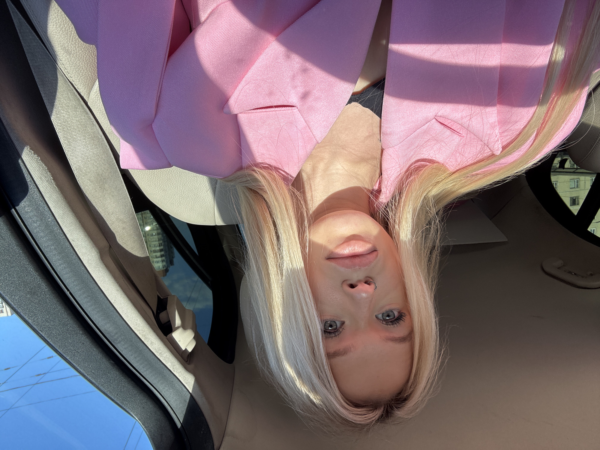 The image shows a young woman with blonde hair and blue eyes, looking at the camera. She is wearing a pink blazer. She is inside a car, with the car's interior visible in the foreground. The window on the left shows a blue sky and some buildings in the distance, suggesting it is daytime. Sunlight is casting shadows on the woman's face and blazer. The image is oriented sideways.