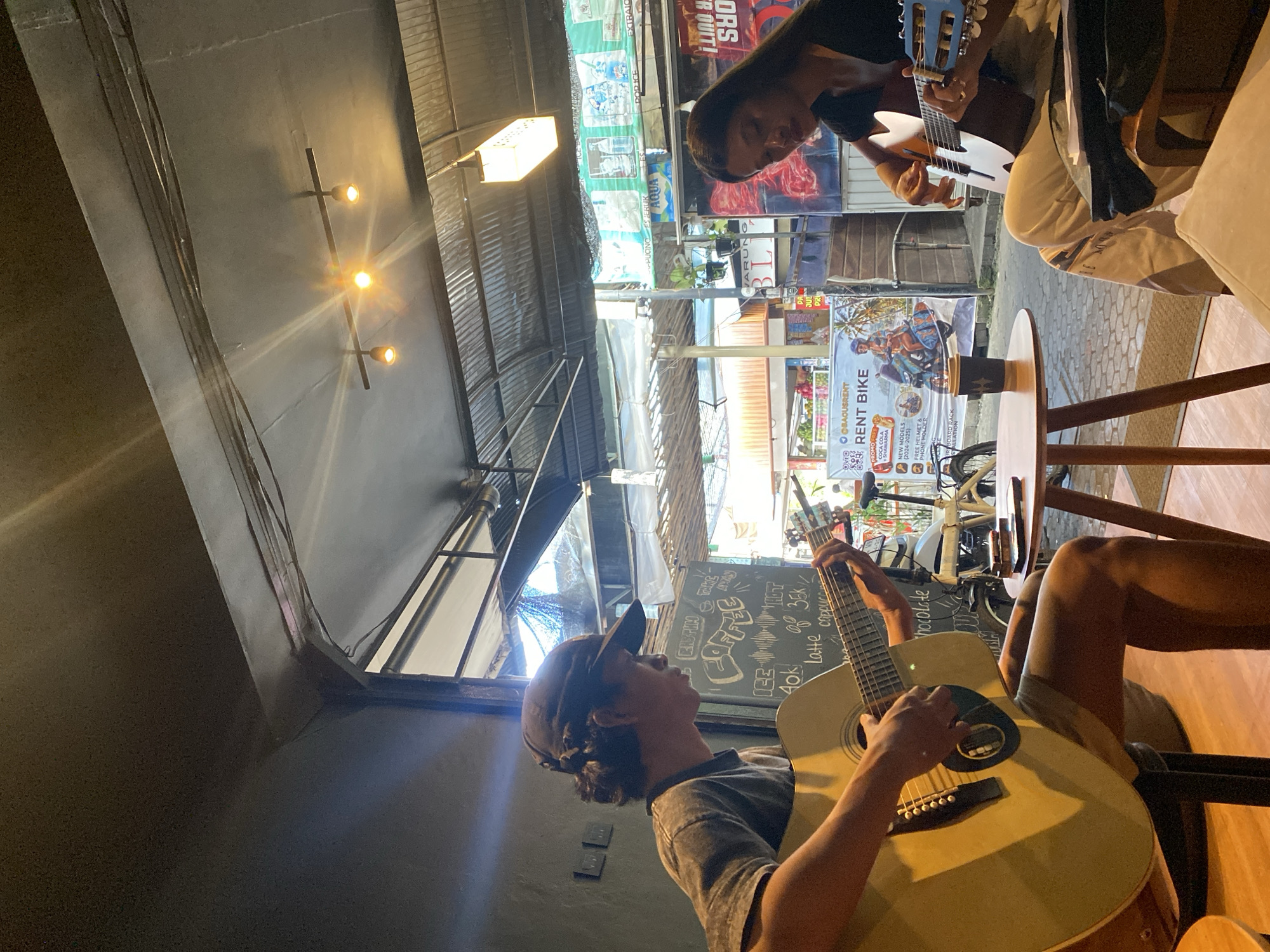 The image shows two young adults playing acoustic guitars in an open-air cafe in Kuta, Indonesia. The person on the left is wearing a cap and playing a large acoustic guitar, while the person on the right is playing a smaller guitar. They are seated at small wooden tables with a coffee cup on one of them. In the background, there are posters for a rent bike service and a menu board with coffee prices. The cafe appears to be located on a street with shops and motorbikes visible. The lighting suggests it is daytime, with sunlight filtering through the corrugated metal roof.