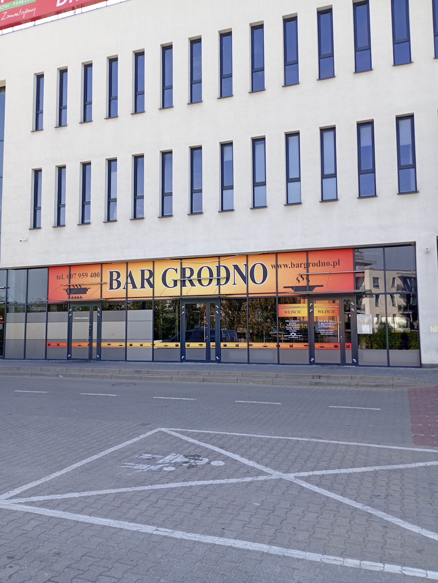 The image shows the facade of a building, likely a restaurant or bar, with a large glass storefront. The sign above the storefront reads "BAR GRODNO" in large, colorful letters, with "www.bargrodno.pl" to the right. Below the sign, there are glass doors and windows. The building appears to be modern, with a white facade and multiple rows of windows. In the foreground, there is a paved area with white lines marking parking spaces, including one designated for people with disabilities, indicated by a wheelchair symbol. The scene is captured during daylight.