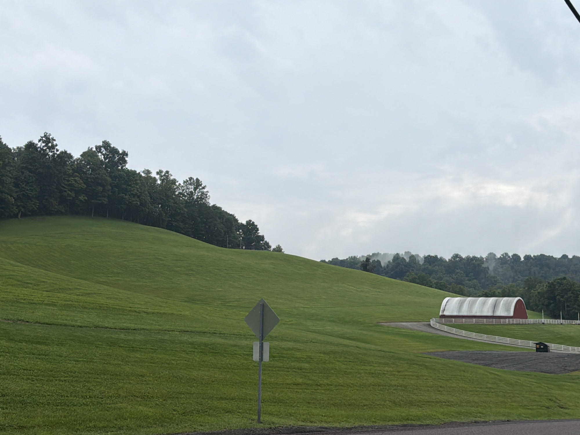 A vast, gently sloping green pasture dominates the foreground of this outdoor, daytime image, under a heavily overcast sky. The pasture exhibits subtle textural variations from mowing patterns, with a narrow, dark strip of what appears to be a road or path curving through it from the right middle ground towards the foreground. A diamond-shaped, muted green traffic sign on a metal pole stands prominently in the left middle ground, with a smaller rectangular sign beneath it.
In the right middle ground, a distinctive red barn with a white, arched roof, resembling a Quonset hut, is situated. A white wooden fence delineates a large area around the barn and extends towards the right edge of the frame. Beyond the barn, a dark, rectangular object, possibly a piece of equipment or a small shed, is visible. The background features a dense line of dark green trees, with a hazy, layered effect suggesting distant hills or mountains veiled by the cloudy weather. The overall environment suggests a rural or agricultural setting in Fallowfield Township, United States of America. There are no people or animals visible in the scene.