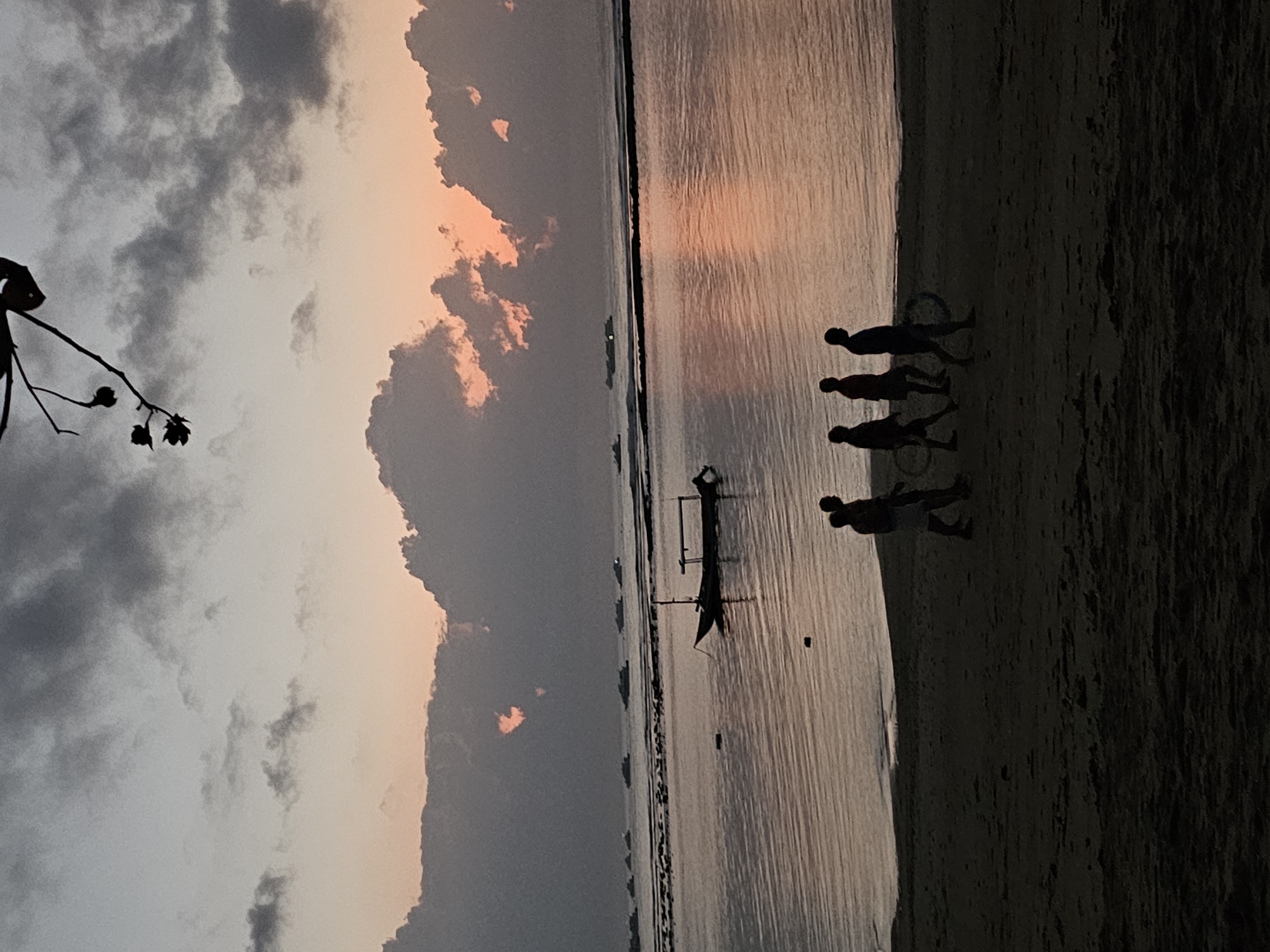 A silhouette of a beach scene at dawn in Denpasar, Indonesia shows a group of four figures walking on the sand in the foreground. One figure appears to be carrying a bicycle wheel. In the middle ground, a traditional Indonesian fishing boat is anchored in calm waters. The background reveals a hazy horizon with several other boats and a soft, pastel-colored sky with dramatic clouds. The overall atmosphere is serene and tranquil, characteristic of a quiet morning by the sea.
