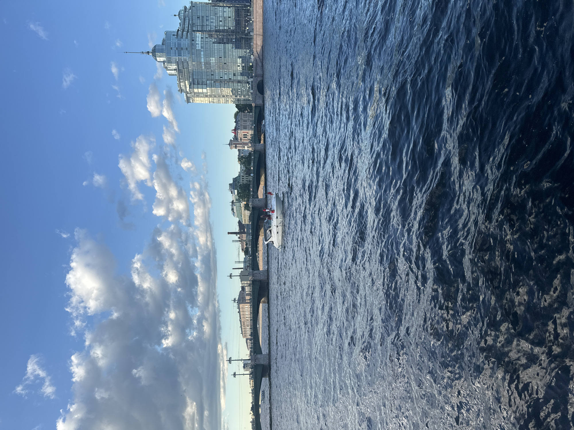The image shows a wide, dark blue river in Saint Petersburg, Russia, with a white motorboat in the foreground. The boat has several people on board, some of whom appear to be waving small flags. In the background, a long, green bridge with stone arches spans the river. Beyond the bridge, a modern, glass-encased building with a distinctive domed top dominates the skyline. Other buildings, including some with red roofs, are visible along the riverbank. The sky is a bright blue with scattered white clouds, suggesting fair weather. The overall scene depicts a view of the city from the water, likely on a sunny day.