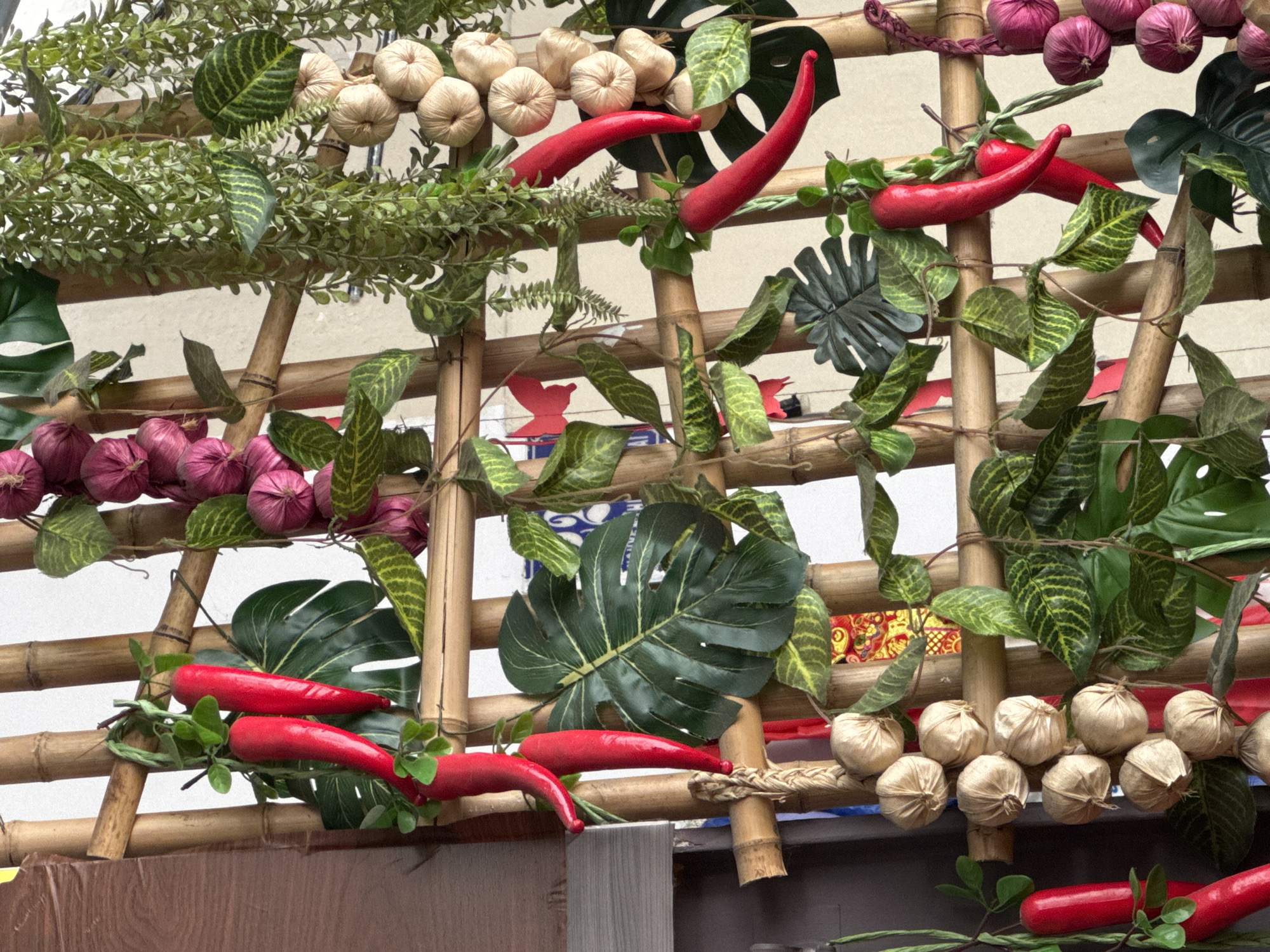 A decorative display of artificial food items is arranged on a bamboo lattice structure, likely in Nha Trang, Vietnam. The arrangement features several bright red chili peppers, strands of small beige-colored bulbs resembling garlic or onions, and bunches of purplish-red bulbs, possibly shallots. Green artificial foliage, including broad monstera-like leaves and trailing vines, is interwoven throughout the bamboo structure. Red paper cutouts of butterflies are also visible. The background is a light-colored wall, and a darker vertical element, possibly a door or furniture, is partially visible on the left. The lighting suggests an indoor or shaded outdoor setting. There are no people or animals present in the image.