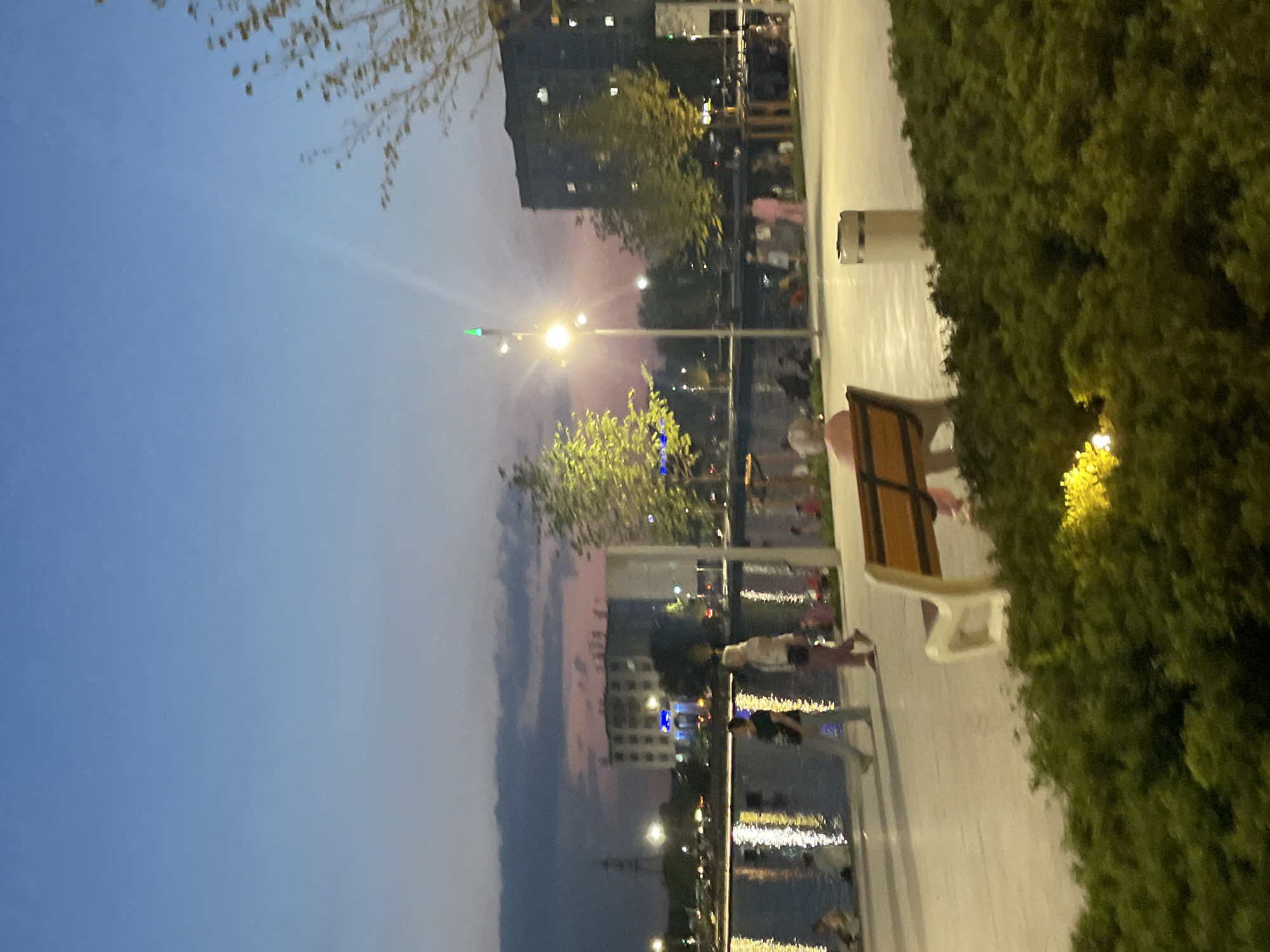 The image depicts a dusk scene at a waterfront promenade in Almaty, Kazakhstan. A paved walkway runs along a body of water, reflecting the ambient light from streetlamps and distant buildings. Several people are visible strolling along the promenade or sitting near the water's edge. In the foreground, a wooden bench is positioned near a patch of dense green bushes, with a trash receptacle next to it. The sky above transitions from a deep blue to lighter shades of pink and orange near the horizon, suggesting the final moments of sunset or the beginning of twilight. The architecture of nearby buildings, including a tall, multi-story structure and what appears to be a tower in the distance, indicates an urban setting. The overall atmosphere is peaceful and recreational.