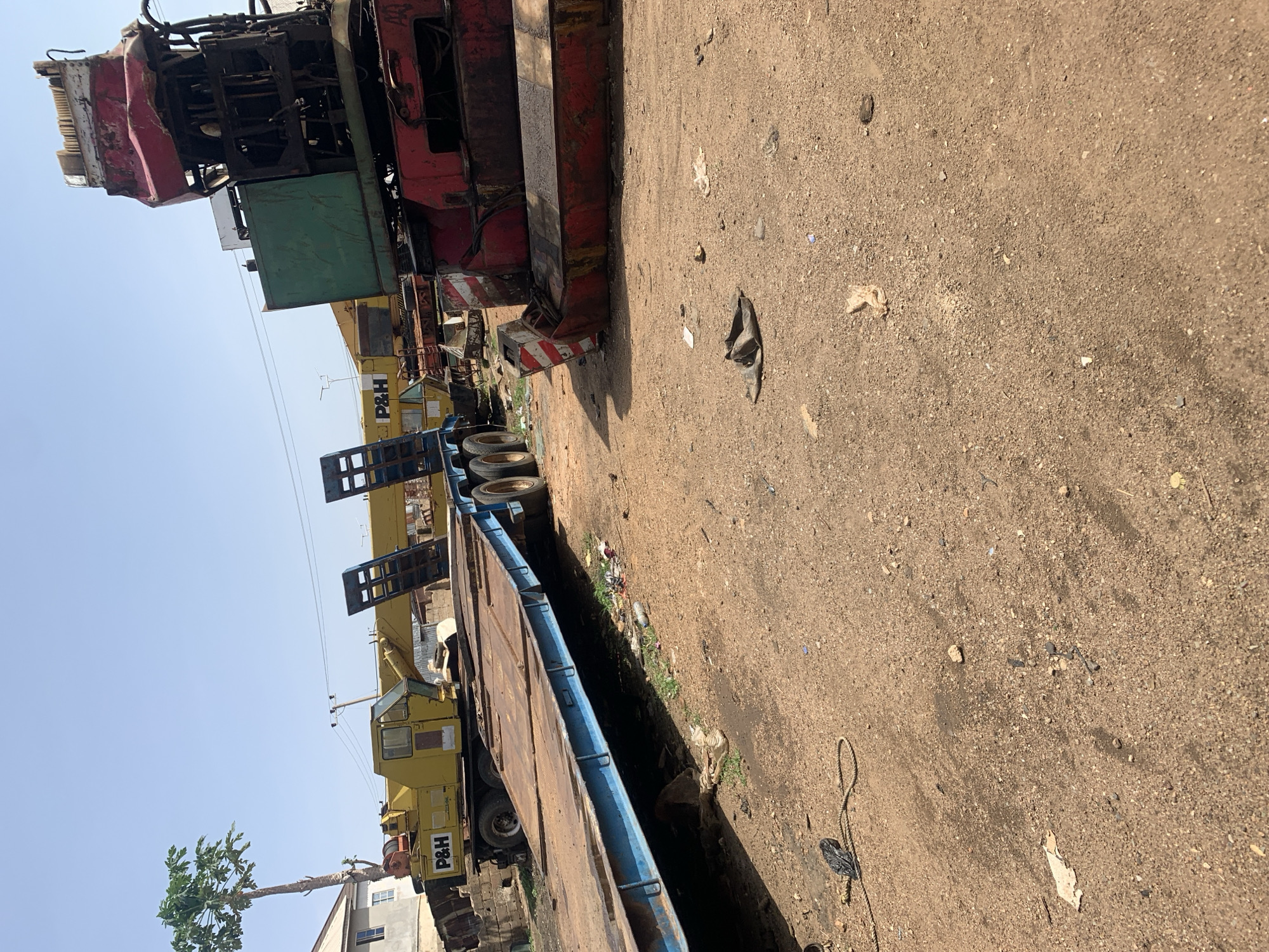 The image captures an outdoor scene at what appears to be an industrial or equipment yard in Rafin Par, Nigeria, under a clear, bright blue sky. The ground is unpaved, consisting of dirt and sand, with scattered debris including small rocks, plastic, and discarded items.
On the left side of the frame, several large pieces of heavy machinery are parked. A prominent red and black heavy-duty crane or construction vehicle with complex mechanical components is visible, partially obscured, its robust structure showing signs of use. Below it, a yellow crane, clearly marked with "P&H" on its side, is visible, featuring a cabin and a large boom arm extending towards the background. More "P&H" branding is seen on another yellow machine further back.
In the foreground, a long, blue and brown flatbed trailer or heavy transport vehicle dominates the lower right portion of the image, positioned alongside a narrow, unkempt ditch containing some sparse vegetation and trash. Several large tires are visible beneath the machinery and the trailer.
No human activity is depicted; the machinery appears to be stationary or stored. The clear sky and the shadows suggest it is daytime, likely mid-morning or afternoon. Utility lines stretch across the background sky. A tall, slender tree with green leaves stands near a building in the far background on the left. On the dirt ground in the foreground, a black plastic bag and a rope are visible among the other detritus.