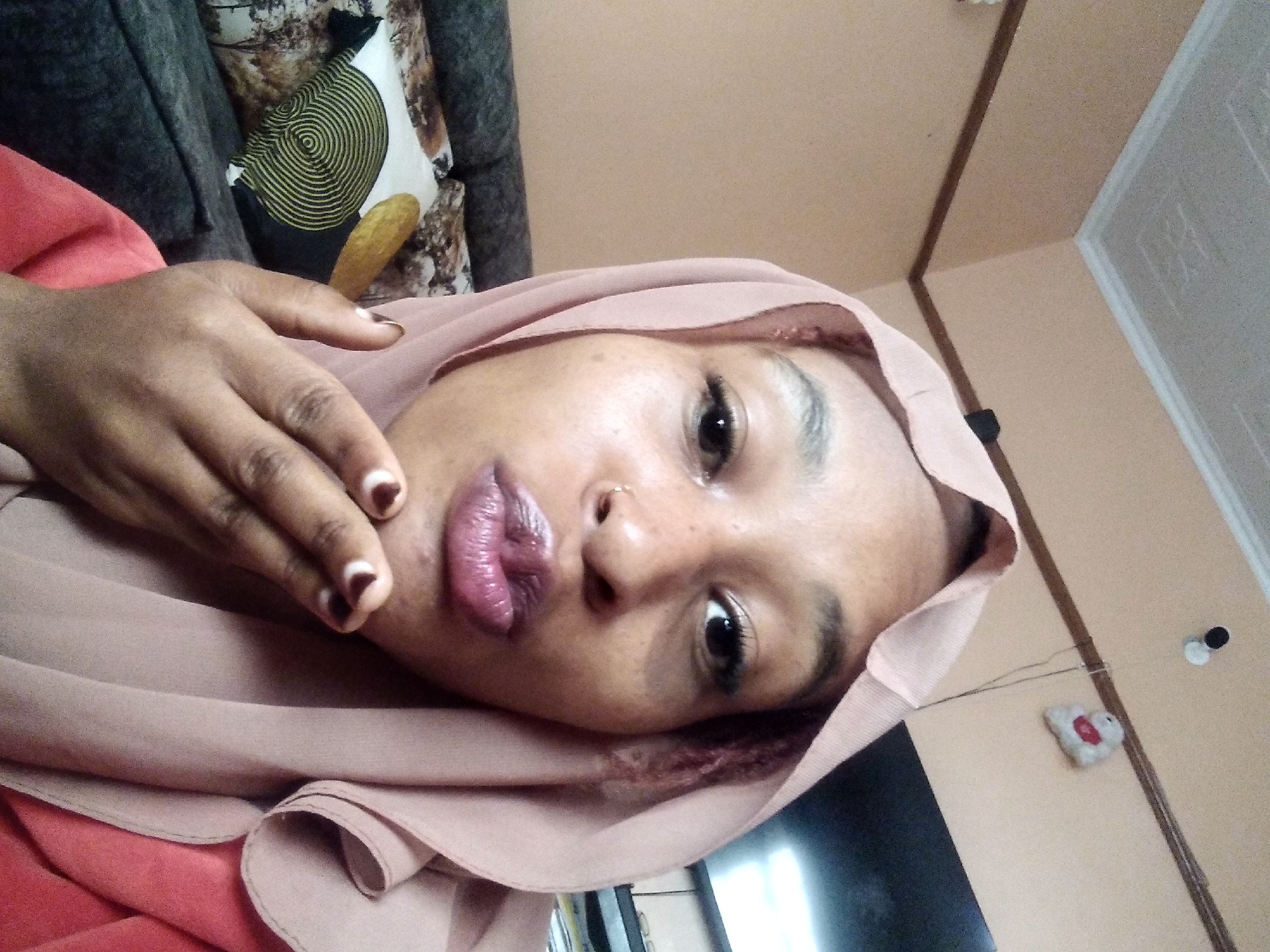 In a domestic indoor setting in Nyeri, Kenya, a young Black woman wearing a pale pink hijab and a coral-colored top is pictured from the chest up. She is leaning forward, facing the camera, and puckering her lips as if blowing a kiss. Her right hand is raised, with fingertips touching her lips. A nose ring is visible on her right nostril. The background shows a cream-colored wall with a white ceiling and a dark, upholstered couch with patterned throw pillows. A television screen is visible in the upper right corner.
