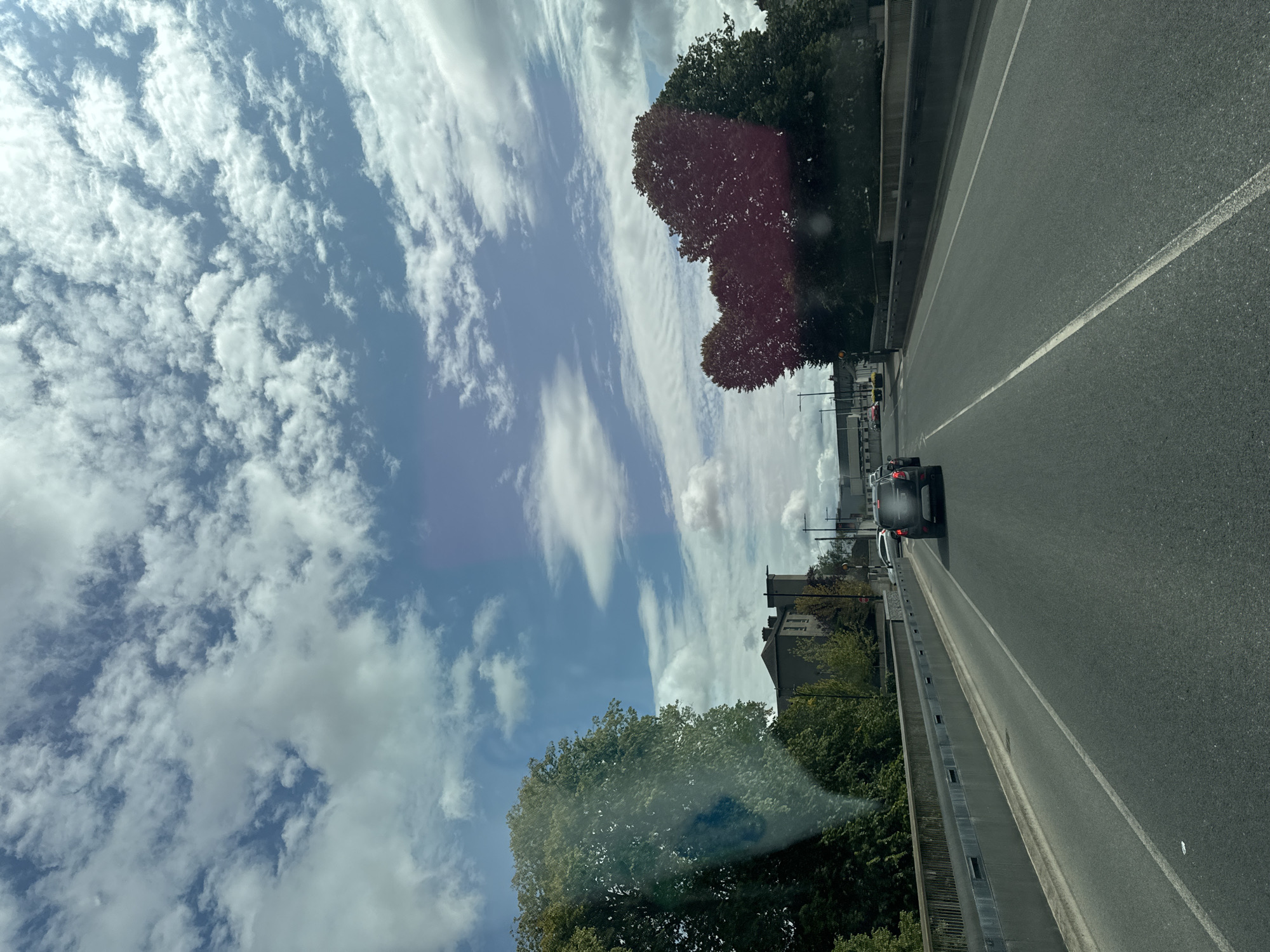 The image depicts a view from a vehicle traveling on a road, likely a bridge, within the city of Kilkenny, Ireland. The sky above is filled with a mix of white and grey clouds against a blue backdrop, suggesting partly cloudy weather.
In the foreground, the asphalt road has clear white lane markings. A concrete barrier with a metal railing runs along the right side of the road, and a similar structure is visible on the left. Several cars are present on the road. In the distance, a black SUV is driving ahead.
On the left side of the road, large, mature trees with green foliage are visible, partially obscuring a building in the background. This building appears to be a multi-story structure with a dark roof and windows. Further ahead, more trees with reddish-brown foliage are prominent on the right side, also framing the view. Beyond the trees, the outlines of other buildings and structures are discernible. Streetlights are also visible along the roadside. The overall impression is an urban or suburban environment with tree-lined roads and architecture consistent with Kilkenny.