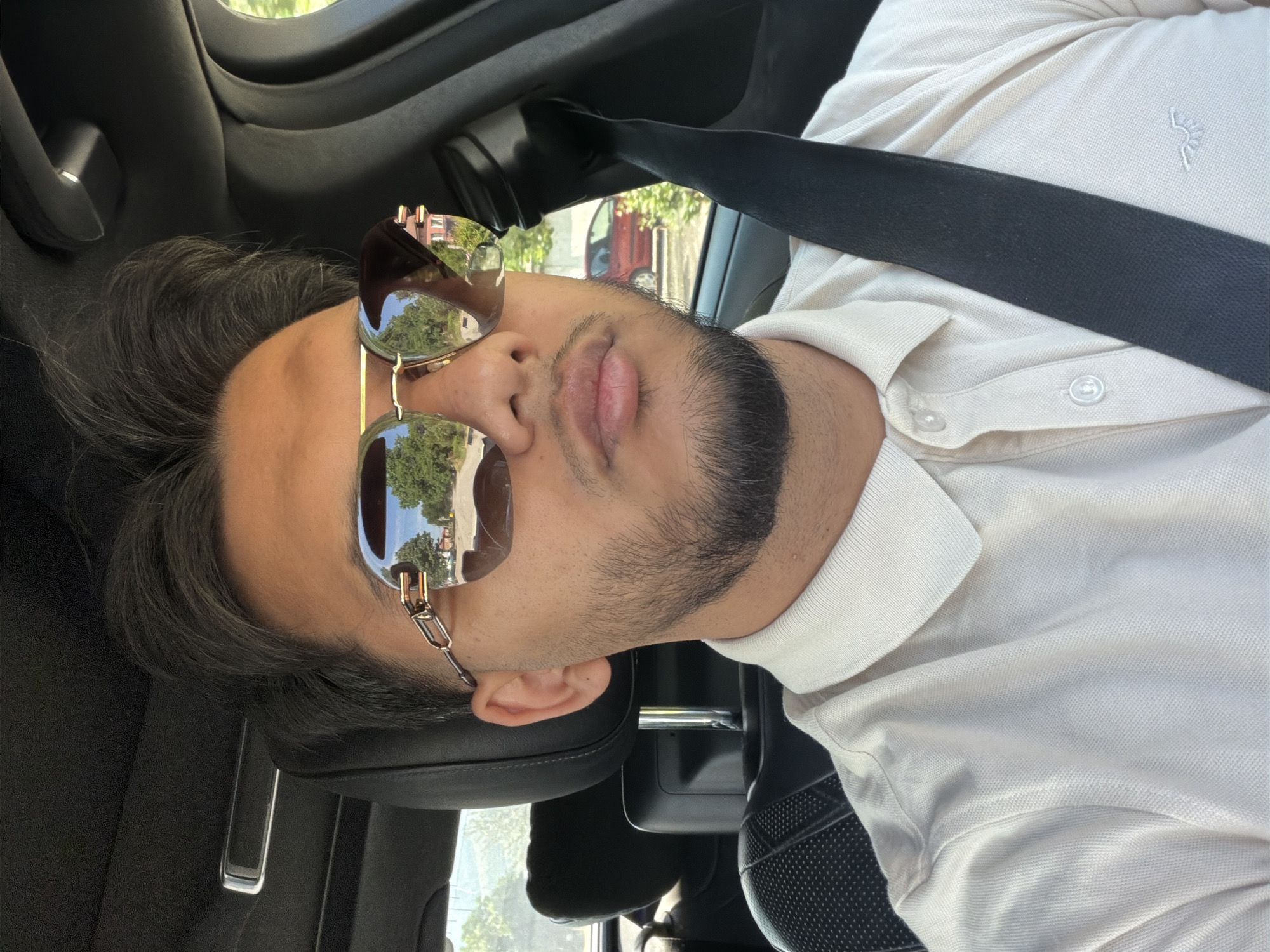 The image presents a close-up, rotated view of a man inside a car, likely a selfie. The subject is a young man with dark hair styled off his forehead and a neatly trimmed beard and mustache. He wears stylish aviator-style sunglasses with reflective, brown-tinted lenses and a gold-colored frame that features distinctive chain-link accents on the temples. His eyes are obscured by the sunglasses, and his lips are slightly parted.
He is dressed in a light beige or off-white short-sleeved polo shirt, complete with a collar and visible buttons down the placket. A small, embroidered logo, appearing to be an abstract plant or tree design, is situated on the left chest of his shirt. A black seatbelt is fastened across his chest, suggesting he is seated in a vehicle.
The setting is the interior of a car, with dark-colored upholstery and elements of the car's interior visible around the man. The reflections in his sunglasses, as well as views through the car windows, clearly indicate an outdoor, daytime environment. These reflections show vibrant green trees, parts of buildings (one appearing to be red brick), and what seems to be a road or street, implying the car is in an urban or suburban setting. The brightness of the reflections suggests sunny or clear weather. The image is oriented with the man's head facing right, indicating a 90-degree clockwise rotation. No text is visible in the image.
