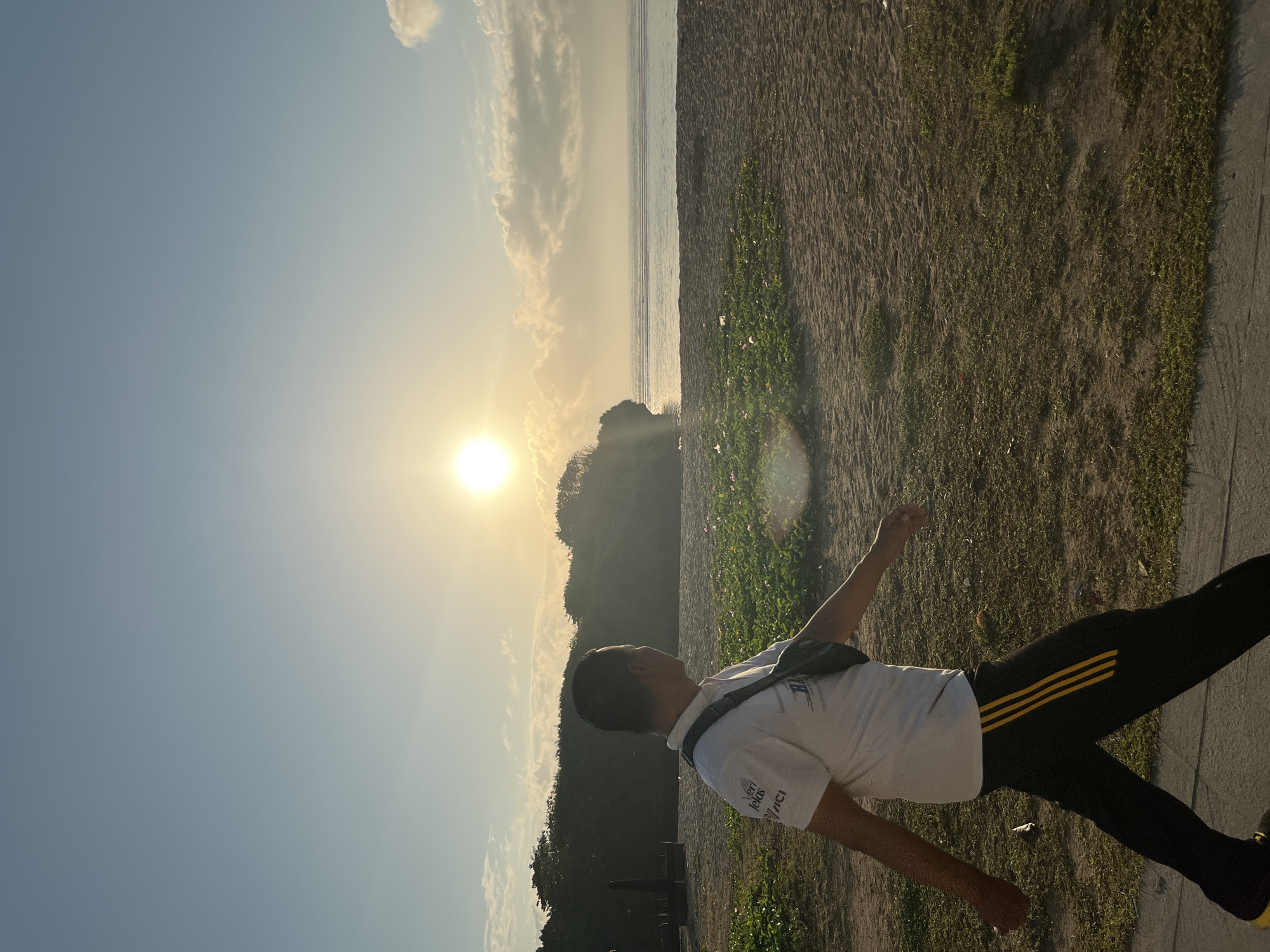 A man with short dark hair, wearing a white polo shirt and dark pants with yellow stripes, walks on a paved path next to a beach. He is seen from the side, facing away from the camera and looking towards the ocean. A dark fanny pack is worn across his chest. The beach sand is visible, with patches of green vegetation and a few scattered pink flowers. The ocean stretches out to the horizon, with gentle waves. In the background, a rocky outcrop covered in trees is visible, with the sun setting or rising behind it. The sky is a soft blue with some scattered clouds, illuminated by the bright sun, which casts a warm glow on the scene. The image appears to be taken in the city of Kuta Selatan, Indonesia, given the coastal environment and the characteristic tropical vegetation.