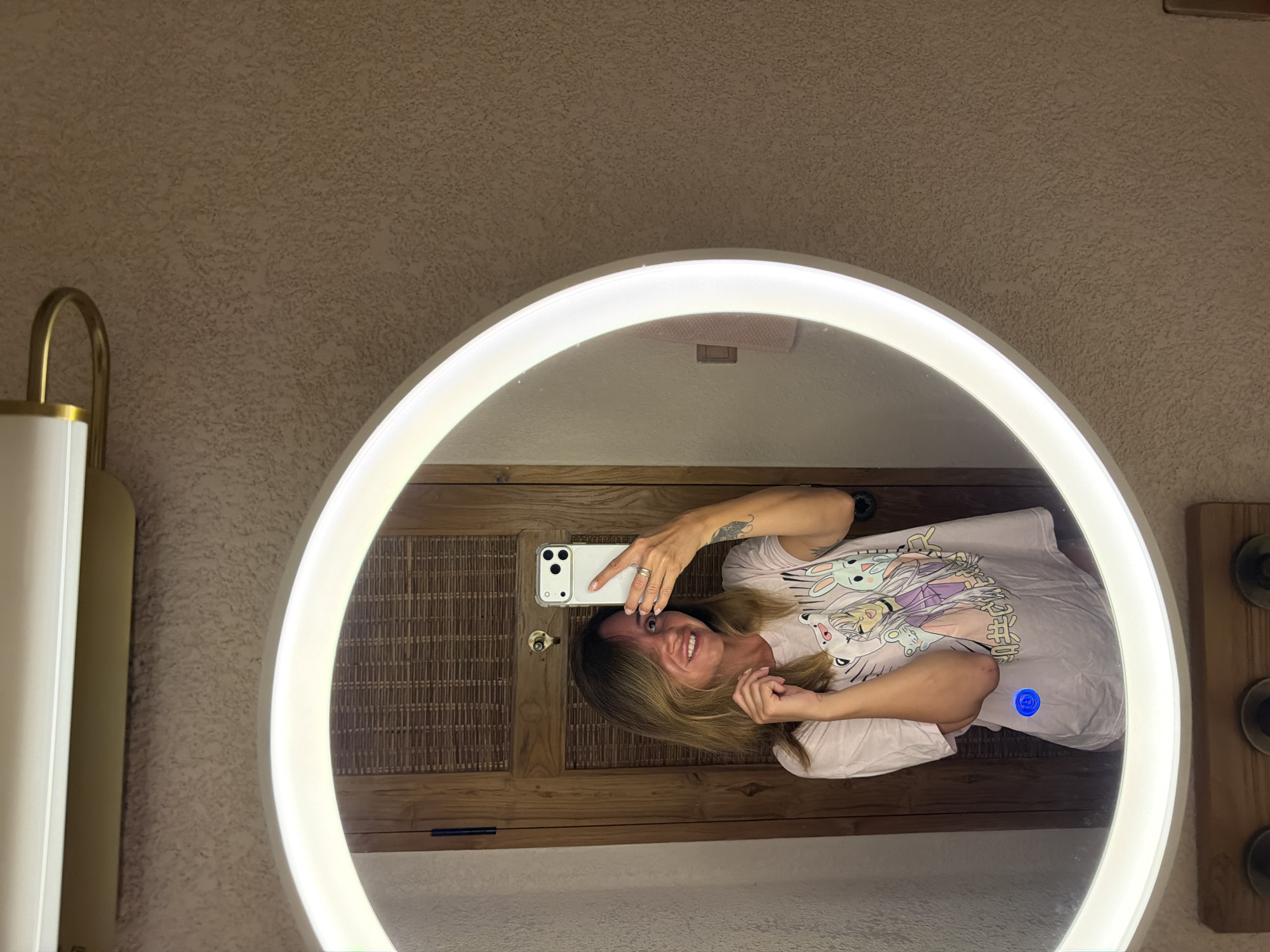 A young woman with blonde hair and a pink t-shirt with anime characters on it is taking a selfie in a circular mirror. The mirror is illuminated by a bright white light ring around its perimeter. Behind the woman, a wooden door with a woven texture is visible. The wall surrounding the mirror has a rough, textured finish. The scene appears to be in a bathroom, possibly in Mengwi, Indonesia.