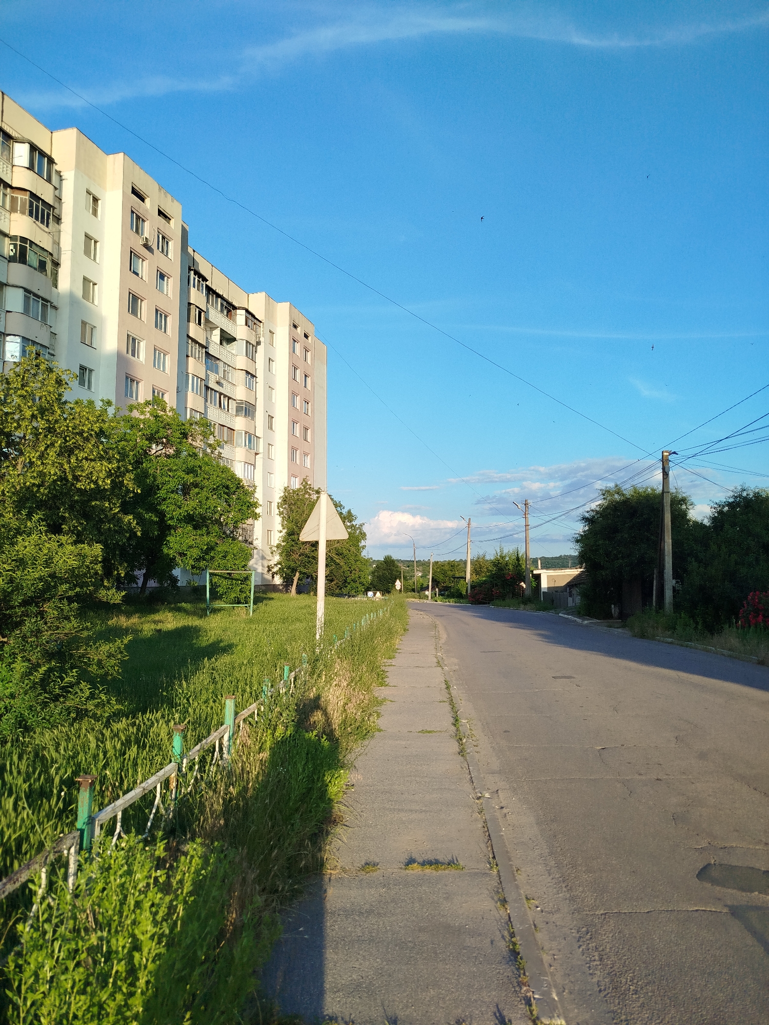 This media file depicts an outdoor scene on a sunny day. The prominent subjects include a tall, multi-story apartment building with numerous windows and balconies, a paved road with a concrete sidewalk, a grassy area with a fence, and several trees. In the distance, there are more trees and some low-lying structures, along with utility poles and power lines crisscrossing the sky. A white, triangular signpost is visible on the grassy side.
The scene appears to be set in a residential area, possibly in a town or a suburban environment. The time of day is likely late afternoon or early evening, indicated by the warm, angled sunlight casting long shadows. The sky is bright blue with a few wispy white clouds, suggesting fair weather. The overall mood is tranquil and ordinary, capturing a slice of everyday life. No people are directly visible in the frame, though a shadow suggesting a person is cast on the grass near the fence. There is no visible text in the image. The elements, such as the apartment building, the road, and the utility infrastructure, suggest a developed, though not necessarily modern, urban or semi-urban setting. The presence of a playground-like structure in the grassy area hints at a community space.