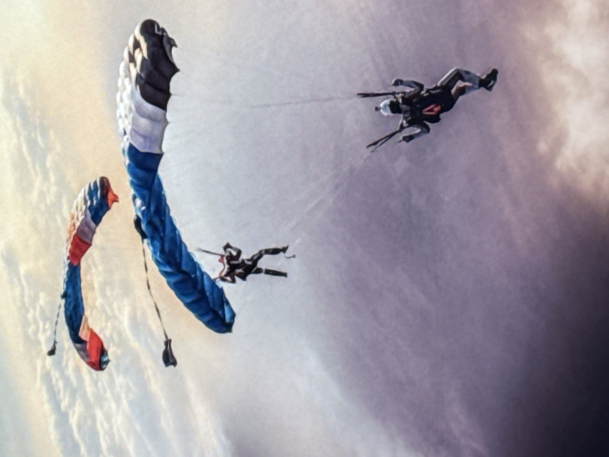 Two skydivers are descending in the sky. One skydiver is wearing a black and grey suit with red accents and a white helmet, and is positioned in the lower right of the frame. The second skydiver is wearing a black suit and is positioned higher up and to the left of the first. Both skydivers have opened parachutes. The parachute above the lower skydiver is blue, white, and black, while the parachute above the higher skydiver is red and blue. The background consists of clouds with a soft, diffused light, suggesting either sunrise or sunset. The image does not contain any identifiable landmarks or features that would place it in Norilsk, Russia.