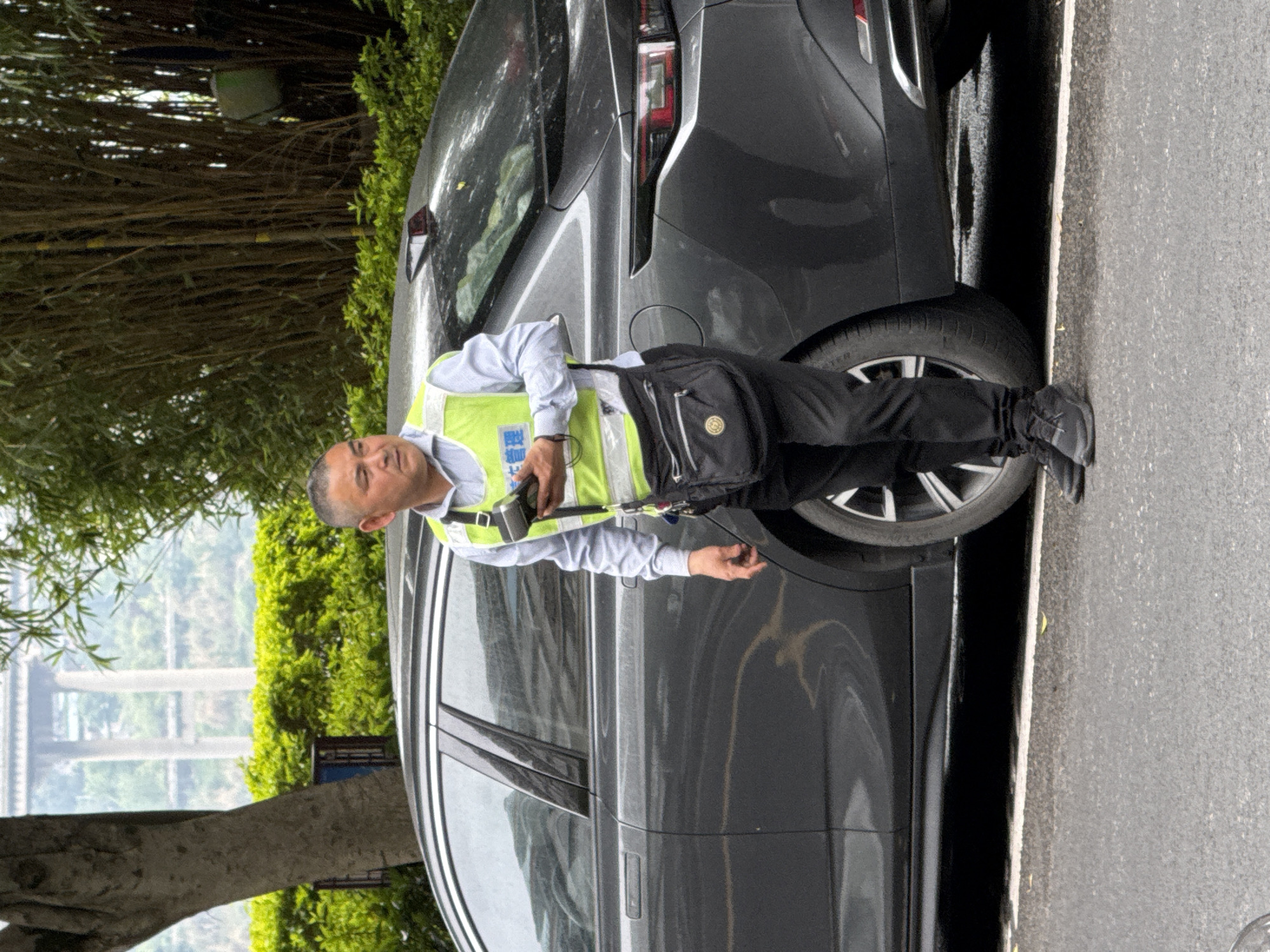 A man wearing a light blue shirt, black pants, and a yellow traffic vest stands next to a parked gray SUV. The man has a black bag slung across his body and holds a small black object in his hand. The scene appears to be on a roadside next to a grove of trees, with the distant structure of a bridge visible. The location is inferred as Jiangjin, China, based on the information provided.