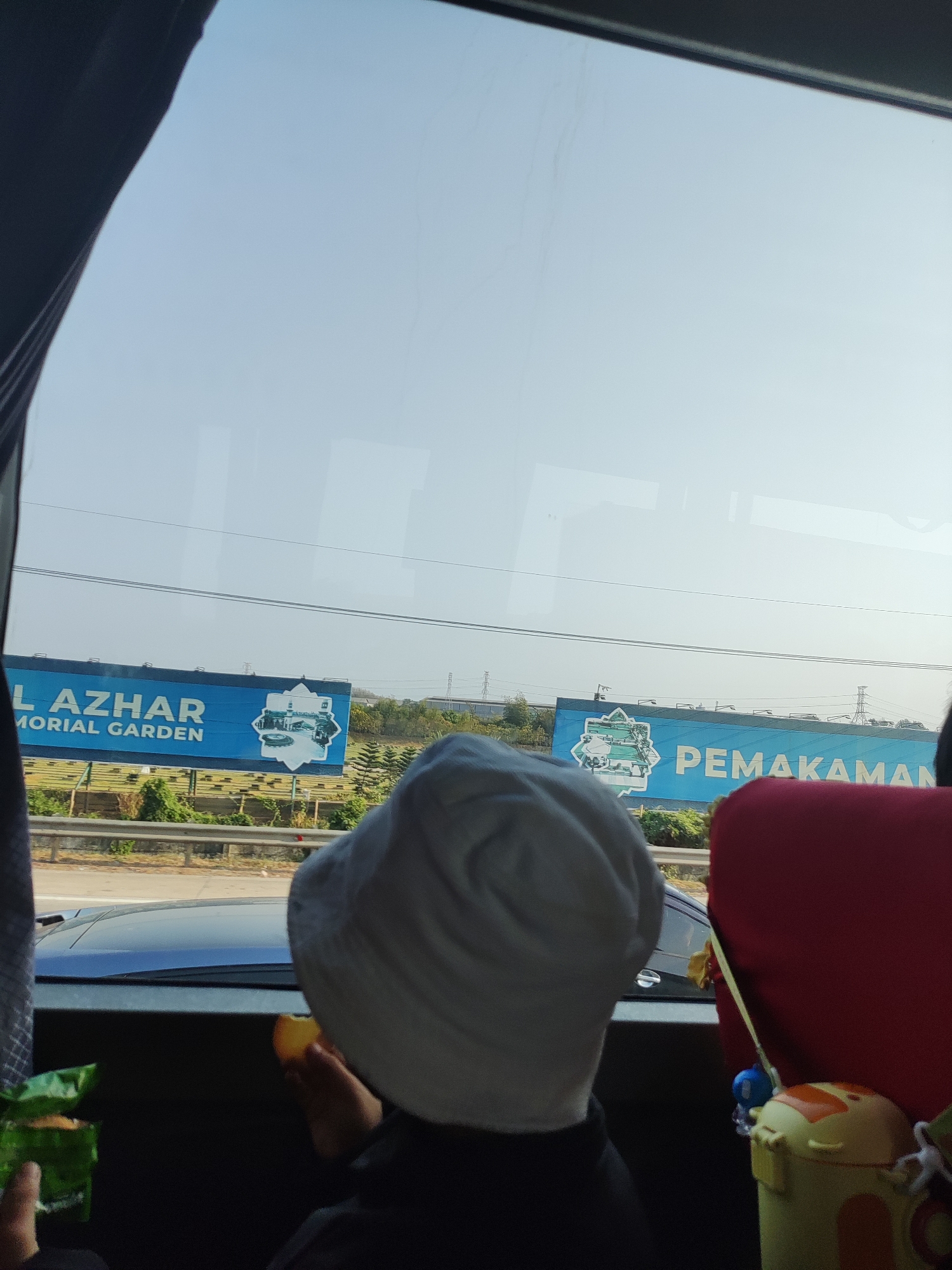 The image shows a view from inside a vehicle, looking out a window. A person wearing a white bucket hat is seated in the foreground, facing away from the camera and holding a piece of fruit. To the left, a green bag of snacks is visible. Through the window, two large billboards are seen. The left billboard reads "AL AZHAR MEMORIAL GARDEN" in white text on a blue background, with an image of a building and a circular water feature. The right billboard displays "PEMAKAMAN" in large white text on a blue background, also featuring a similar graphic. In the distance, there are trees, power lines, and a highway. The sky is clear and blue. The scene is likely in Karawang, Indonesia, given the billboards advertising a cemetery, which is a common sight in populated areas.