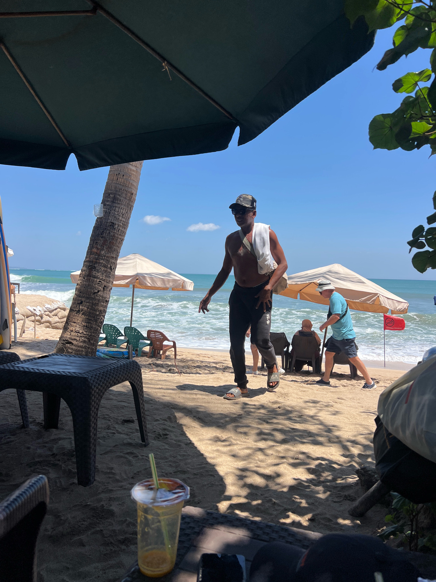 An eye-level, medium outdoor shot captures a bright, sunny day on a sandy beach in Kuta, Indonesia, near the ocean. The foreground is dominated by dark wicker furniture casting distinct shadows across the sand. A clear plastic cup with a yellow-orange beverage and a green straw sits on a table.
In the mid-ground, a man wearing a camouflage baseball cap, sunglasses, and black pants is walking towards the left, a white towel draped over his shoulders. He is barefoot, wearing flip-flops. Behind him, a tan umbrella provides shade, and several beach chairs are visible. Further back, two other men are seated, one wearing a blue t-shirt and shorts, the other in a brown head covering and a seated position. A red flag is planted in the sand near the umbrella. The ocean waves are a prominent feature, with white foam breaking on the shore. A palm tree trunk stands to the left of the frame, and surfboards are propped up against a sandbag barrier on the far left.
The background consists of a clear blue sky with a few scattered white clouds and the vast expanse of the turquoise ocean. Green leaves from a tree are visible in the upper right corner, adding a touch of natural foliage to the scene.