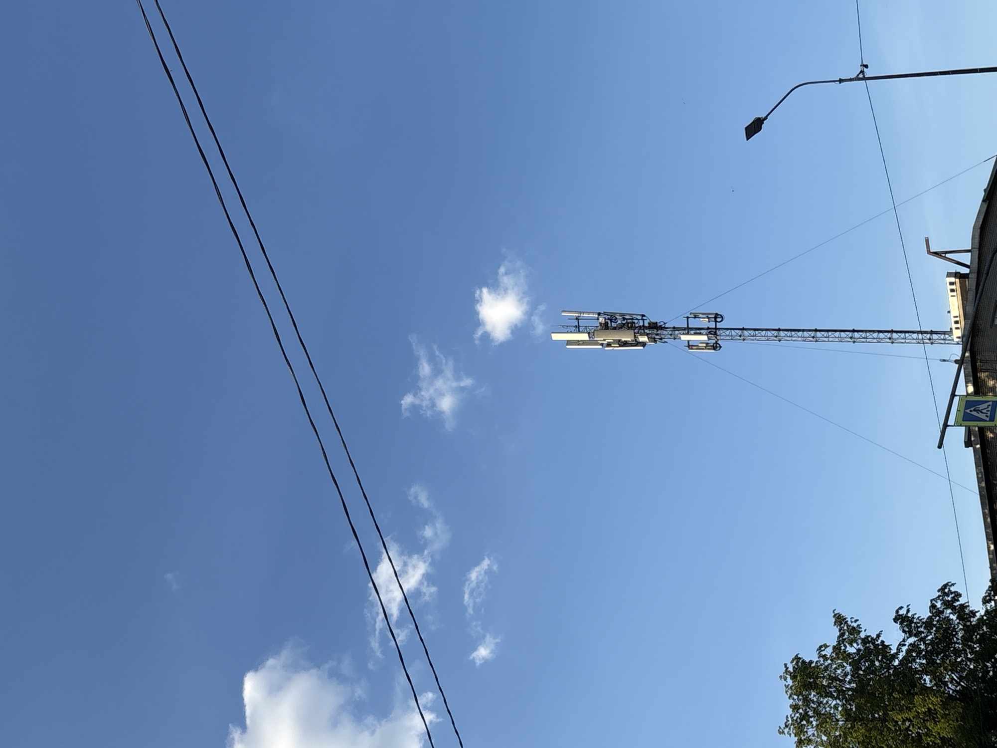 The image presents an upward view of an urban environment against a bright, clear blue sky with a few scattered white cumulus clouds. Dominating the central-right portion of the frame is a tall, slender metal lattice structure, possibly a crane or a communication tower, extending vertically. It is equipped with several horizontal arms bearing rectangular panels, which could be antennas for telecommunications or lighting fixtures.
Two dark, parallel electrical or communication cables stretch diagonally across the upper-left quadrant of the sky. In the upper-right corner, a modern street light with a rectangular head is visible, attached to an arm that extends from a pole. This pole runs down the right edge of the image, revealing part of a multi-story building, possibly constructed of brick or concrete. A pedestrian crossing sign, characterized by a blue square with a white walking figure, is partially visible, hanging near the building. The lower-right corner shows a cluster of vibrant green tree foliage.
The scene depicts a typical daylight setting in an urban area, likely Saint Petersburg, Russia, given the provided context. The weather appears fair and sunny. No people are visible, and there is no discernible activity or event taking place, presenting a static snapshot of city infrastructure against the sky. No explicit text is visible beyond the symbolic pedestrian crossing sign.