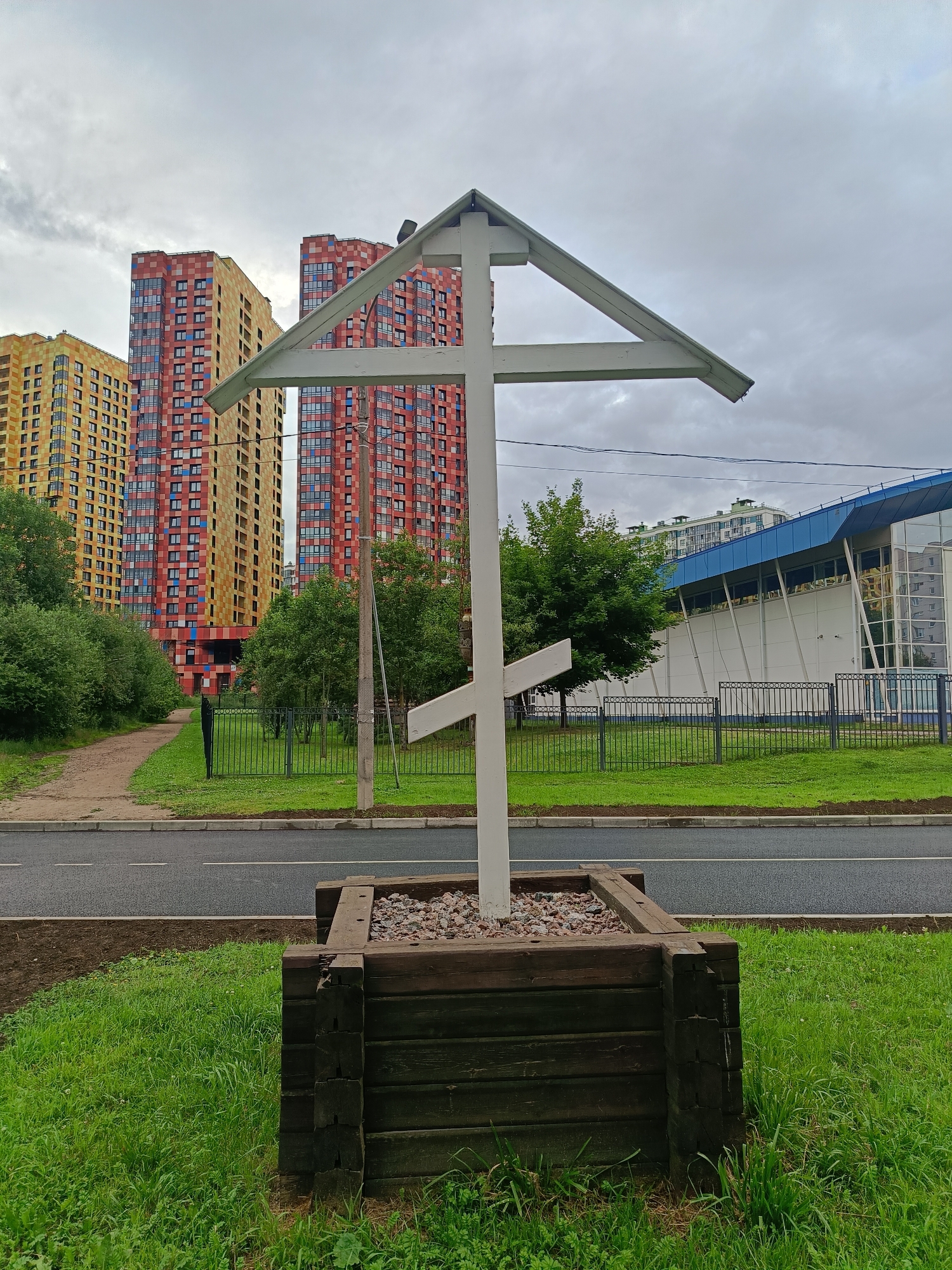 A white wooden Orthodox cross stands in a square wooden planter filled with gravel. The cross has a triangular canopy at the top and a smaller crossbar. In the background, a modern residential complex with bright yellow and red buildings is visible, along with a blue and white building that appears to be a sports complex. A paved road runs in front of the planter, with a grassy area and trees on the other side of the road. The sky is overcast. The scene is located in the city of Saint Petersburg, Russia, specifically in the residential area of Murino. There are no people in the image.