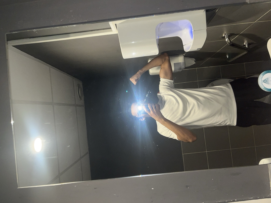 The image displays a male individual taking a mirror selfie in a public restroom. The exact location within Strasbourg, France, is not visually identifiable. The subject, visible from the mid-torso upward, is wearing a white t-shirt and dark trousers. He is positioned with his right arm bent, flexing his bicep. His face is obscured by a bright flash from the camera or phone he is holding. The restroom environment, reflected in the mirror, consists of dark grey tiled walls, a white drop ceiling with square panels, a circular ceiling light, and a circular ventilation grate. On the right side of the reflection, a white automatic hand dryer with an active blue light is mounted to the wall, positioned above a chrome grab bar. A white wall-mounted soap or sanitizer dispenser is visible to the left of the hand dryer, with a portion of a white toilet bowl visible beneath it. A circular sticker featuring "AUTOMATIC" text and a pyramid graphic is present in the lower right quadrant of the mirror's reflection.