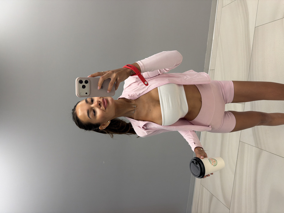 A woman in workout attire takes a selfie. She wears a white top and light pink shorts with a matching jacket. In her left hand, she holds a coffee cup. Her hair is pulled back in a ponytail. The background is a gray wall and light tile floor. She has a red wristband on her left arm and a silver necklace. She is holding a light-colored smartphone with multiple cameras in front of her face.