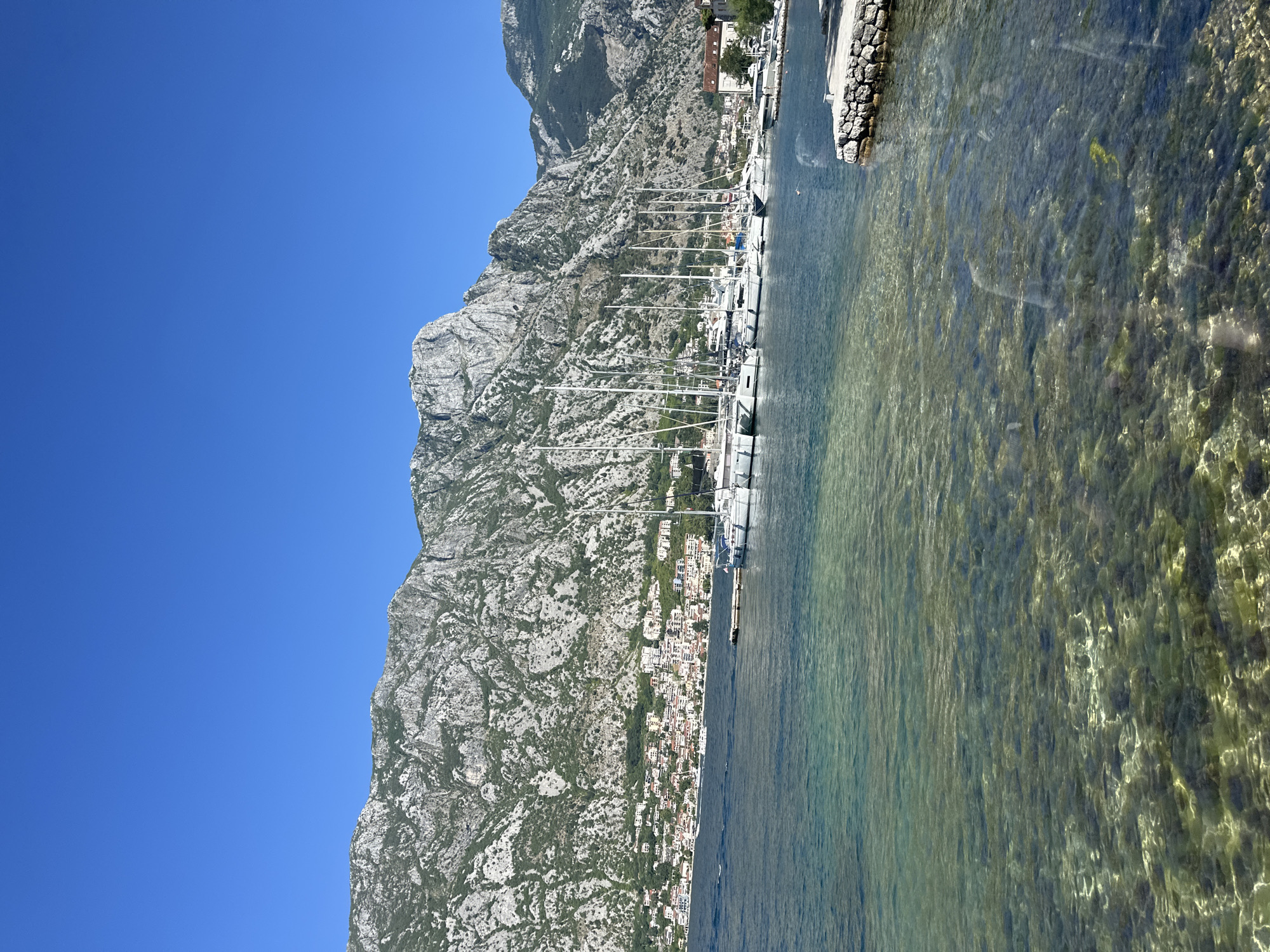 The image shows a clear, sunny day with a bright blue sky above a bay in Prčanj, Montenegro. In the foreground, the shallow, clear water allows visibility of the rocky seabed. A stone walkway juts out into the water on the right side of the frame. Further out in the bay, a line of white sailboats are docked along a pier. Behind the boats, a town with buildings of various sizes and colors stretches along the coastline, backed by steep, green and rocky mountains that dominate the background. The overall scene depicts a tranquil coastal landscape.