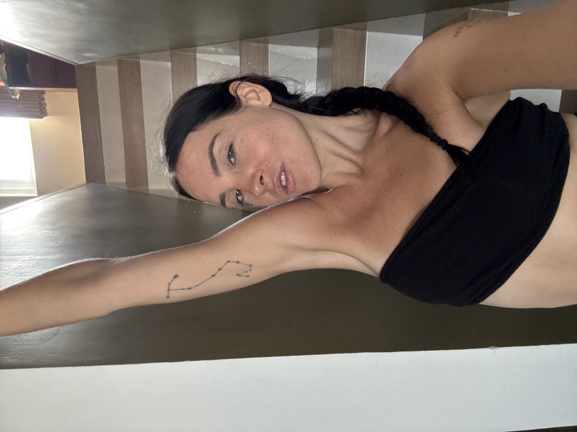 In the city of North Kuta, Indonesia, a woman with dark hair braided to her side and a black strapless top is captured in a close-up selfie. Her right arm is raised above her head, revealing a tattoo on her upper arm. She is positioned in front of a staircase with light brown steps and a dark brown railing. The background is dimly lit, with a window visible at the top of the stairs letting in natural light. The woman's facial expression is neutral, with her head slightly tilted.