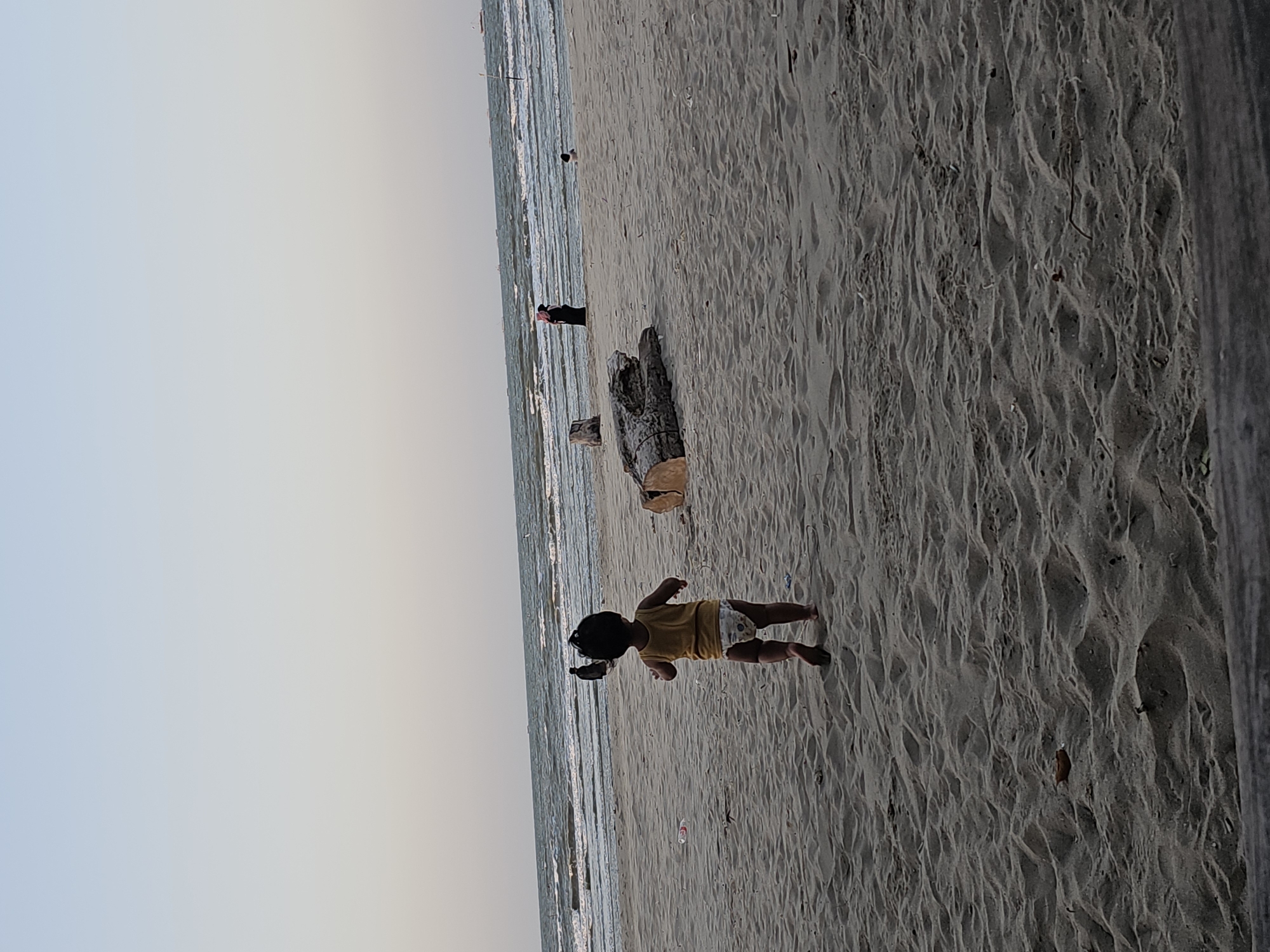 In Dalegan, Indonesia, a toddler wearing a yellow tank top and a diaper runs across a sandy beach towards the ocean. The child's back is to the viewer. In the distance, several people are visible near the water's edge. Two figures stand together, one in dark clothing and the other in a red head covering. Further out, a small child sits in the water. The ocean has moderate waves. Large pieces of driftwood are scattered on the sand. The sky is a pale gradient of blue and white. The scene suggests a casual day at the beach.