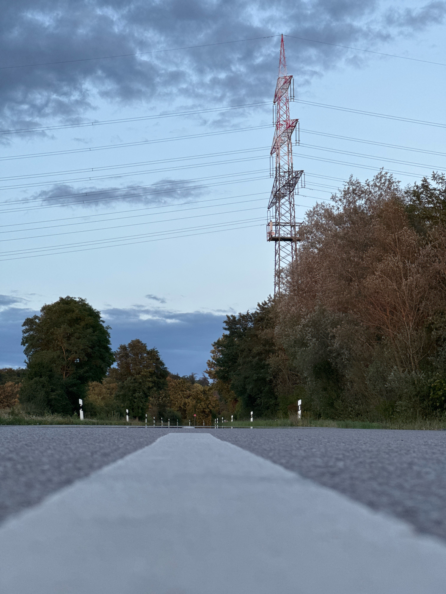 The image depicts a low-angle view of a road with a prominent white line in the foreground, extending towards a rural landscape. In the middle ground, a line of trees and foliage forms a natural barrier. Dominating the background is a tall, red and white lattice electricity pylon, with power lines stretching across the sky. The sky is overcast with a mix of blue and grey clouds. The scene appears to be located near the town of Wettstetten, Germany, indicated by the presence of typical German road markers and the overall rural aesthetic. No people or vehicles are visible in the image.