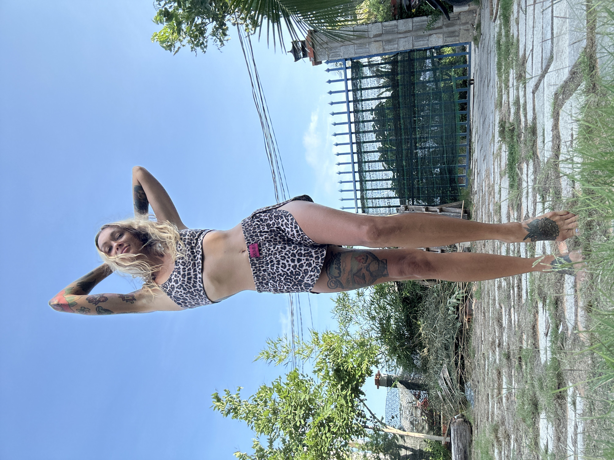 A woman with blonde hair and multiple tattoos stands outdoors in what appears to be a backyard. She is wearing a leopard-print bikini top and matching shorts. Her hands are behind her head, and she is looking towards the camera with a slight smile. The background features a blue sky, lush green trees, a metal fence, and a paved pathway. The scene suggests a warm, sunny day in a residential area, possibly Mũi Né, Vietnam, given the vegetation and architectural style of the fence and gate.