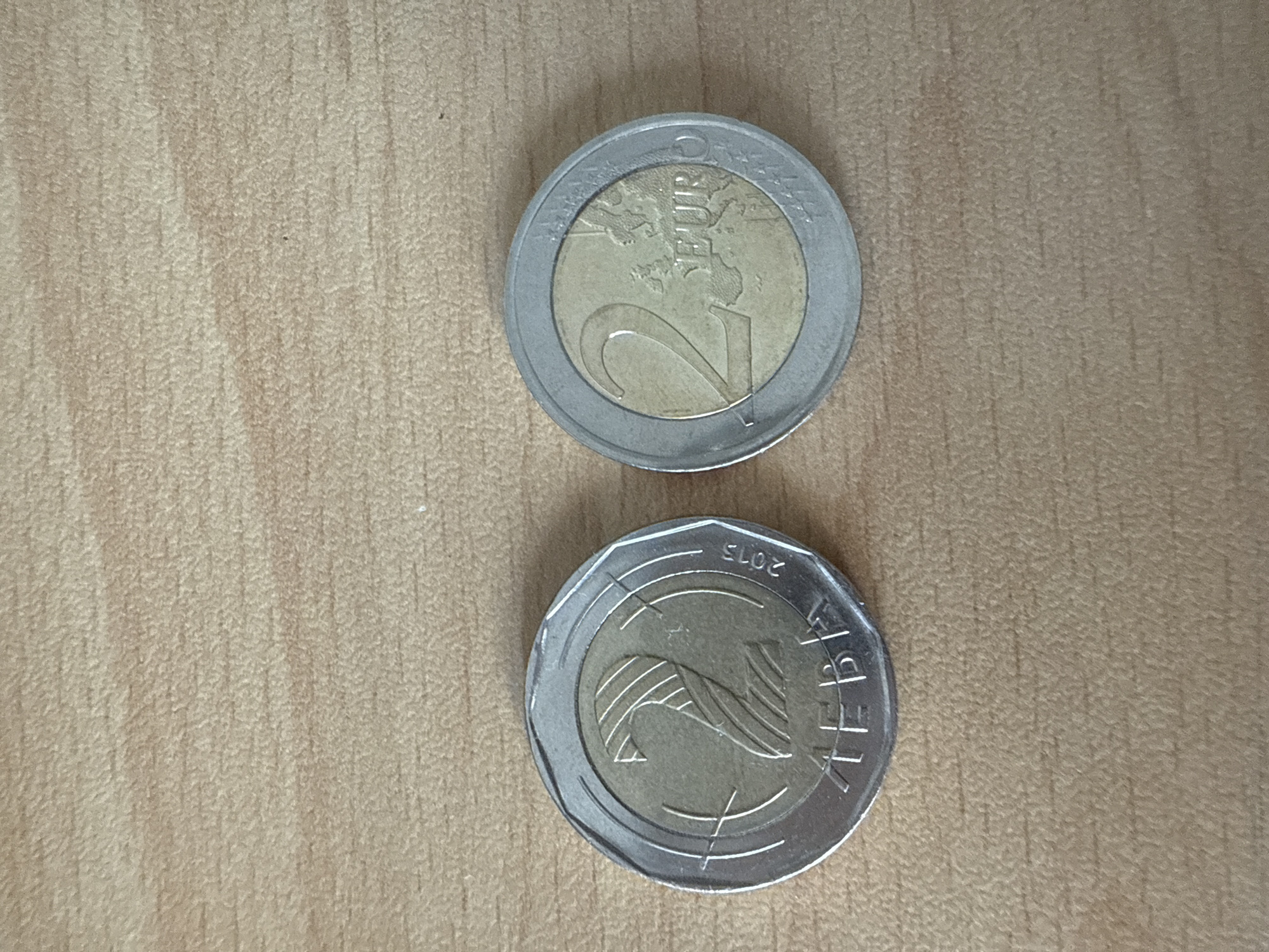 The image shows two 2 euro coins on a wooden surface. The first coin displays the standard "2 EURO" text and a map of Europe. The second coin appears to be a commemorative edition. The text "РЕПУБЛИКА БЪЛГАРИЯ" (Republic of Bulgaria) and the year "2015" are visible on the coin. This edition may have originated in Bulgaria, although that cannot be said with absolute certainty. The location in Varna, Bulgaria is unconfirmed.