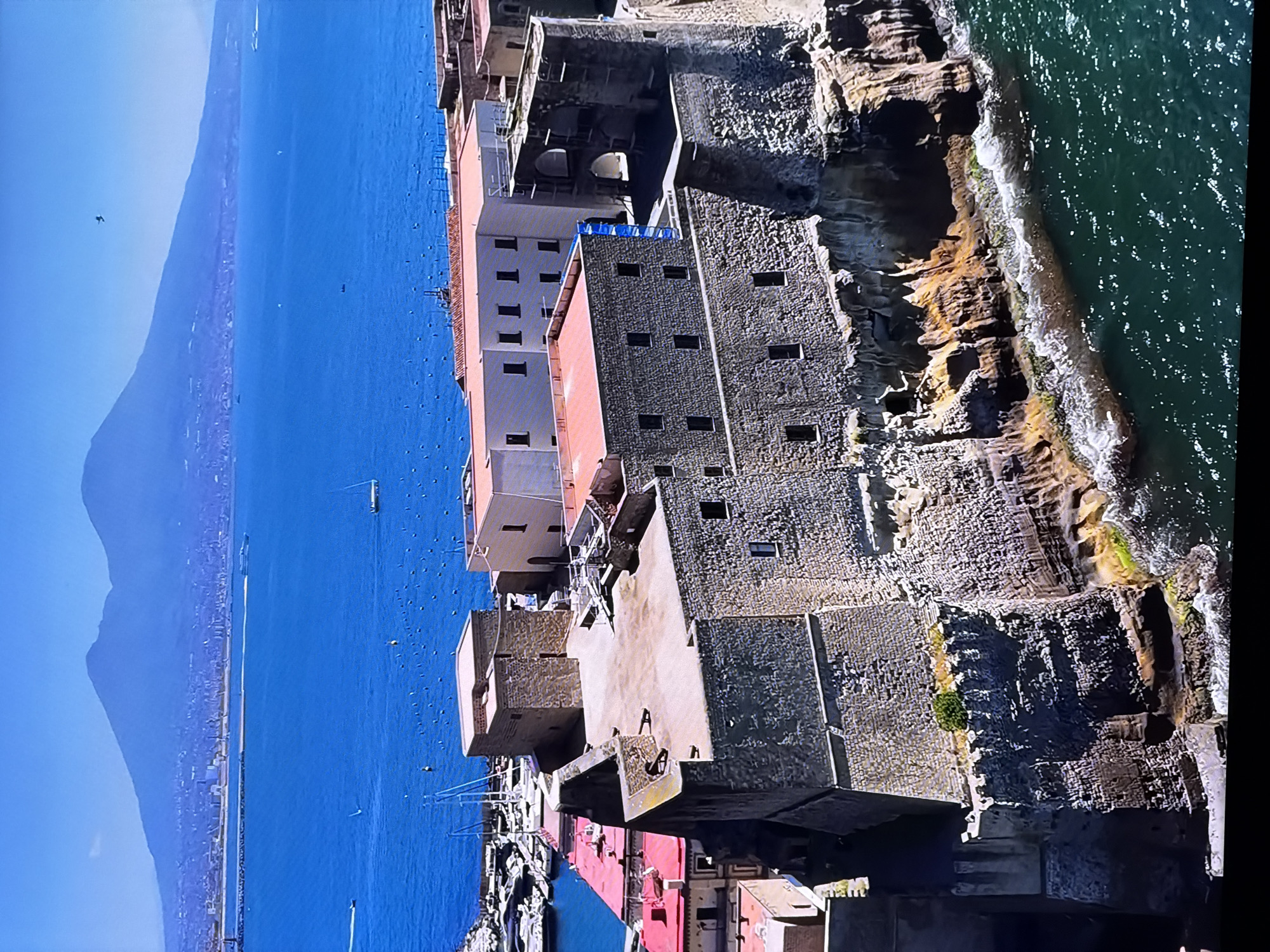 The image depicts a coastal view of a historic castle with a prominent mountain range in the background. The castle, constructed of stone, features crenellated walls and towers, suggesting a fortification. It sits on a rocky cliff overlooking a vibrant blue body of water. Several boats are visible in the water near a harbor, with masts indicating sailboats. In the distance, a city skyline stretches along the coast, leading up to the imposing mountainous landscape. The sky is clear and blue, suggesting fair weather.
This scene is located in Naples, Italy, at Castel dell'Ovo, with Mount Vesuvius in the background. The prompt's assertion that this location is the city of Chirchik, Uzbekistan, is incorrect.