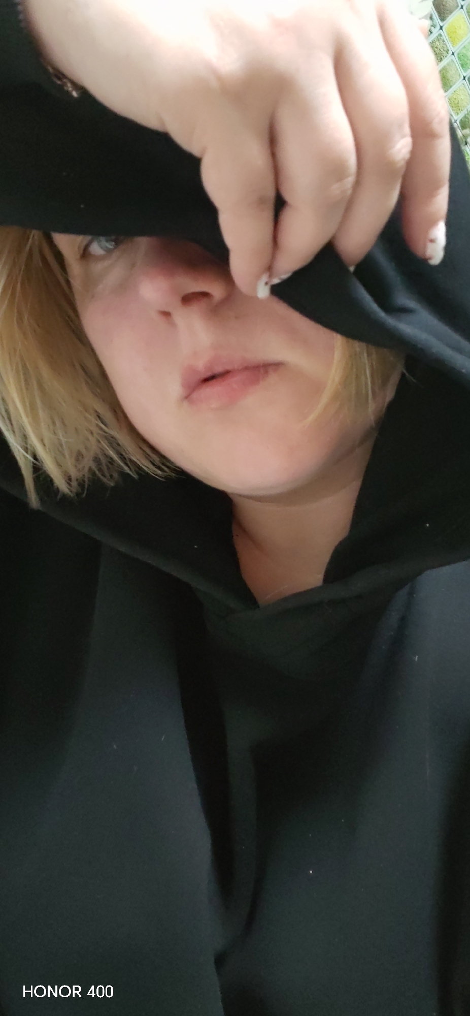 A person with blond hair and fair skin is wearing a black hooded sweatshirt, with the hood pulled up and partially covering the face. One hand is holding the hood fabric near the forehead. The visible fingernails on the hand are painted white with small, dark spots. The person has blue eyes. The environment appears to be indoors. Text "HONOR 400" is in the lower left corner.