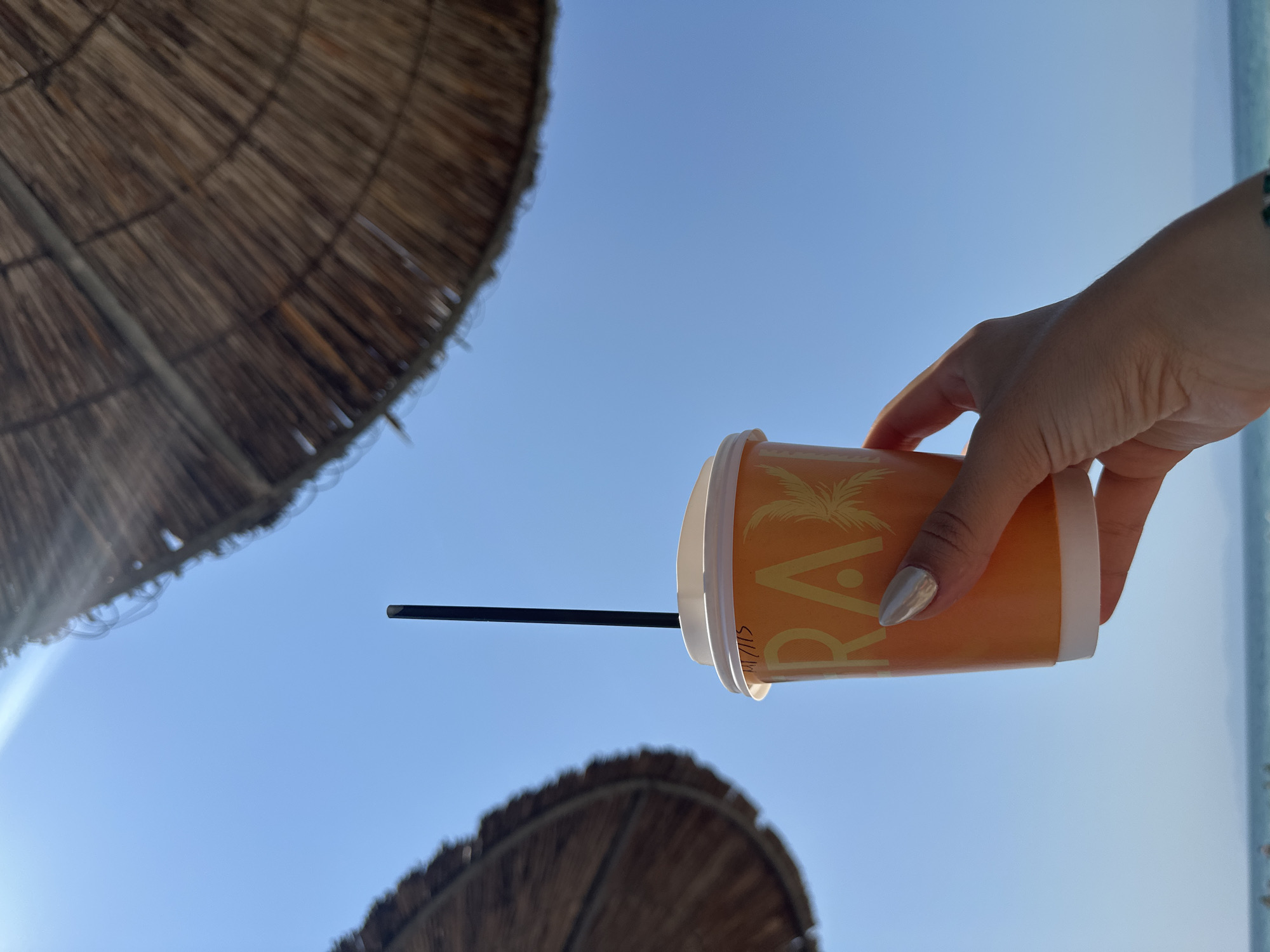 A person's hand, with silver nail polish, holds an orange paper cup with a white lid and a black straw. The cup has a palm tree graphic and the word "RAX" printed in yellow. The background is a clear blue sky with two straw-covered beach umbrellas visible at the top. The scene appears to be at Ofrynio Beach, Greece, with the sea and distant land visible at the bottom of the frame.